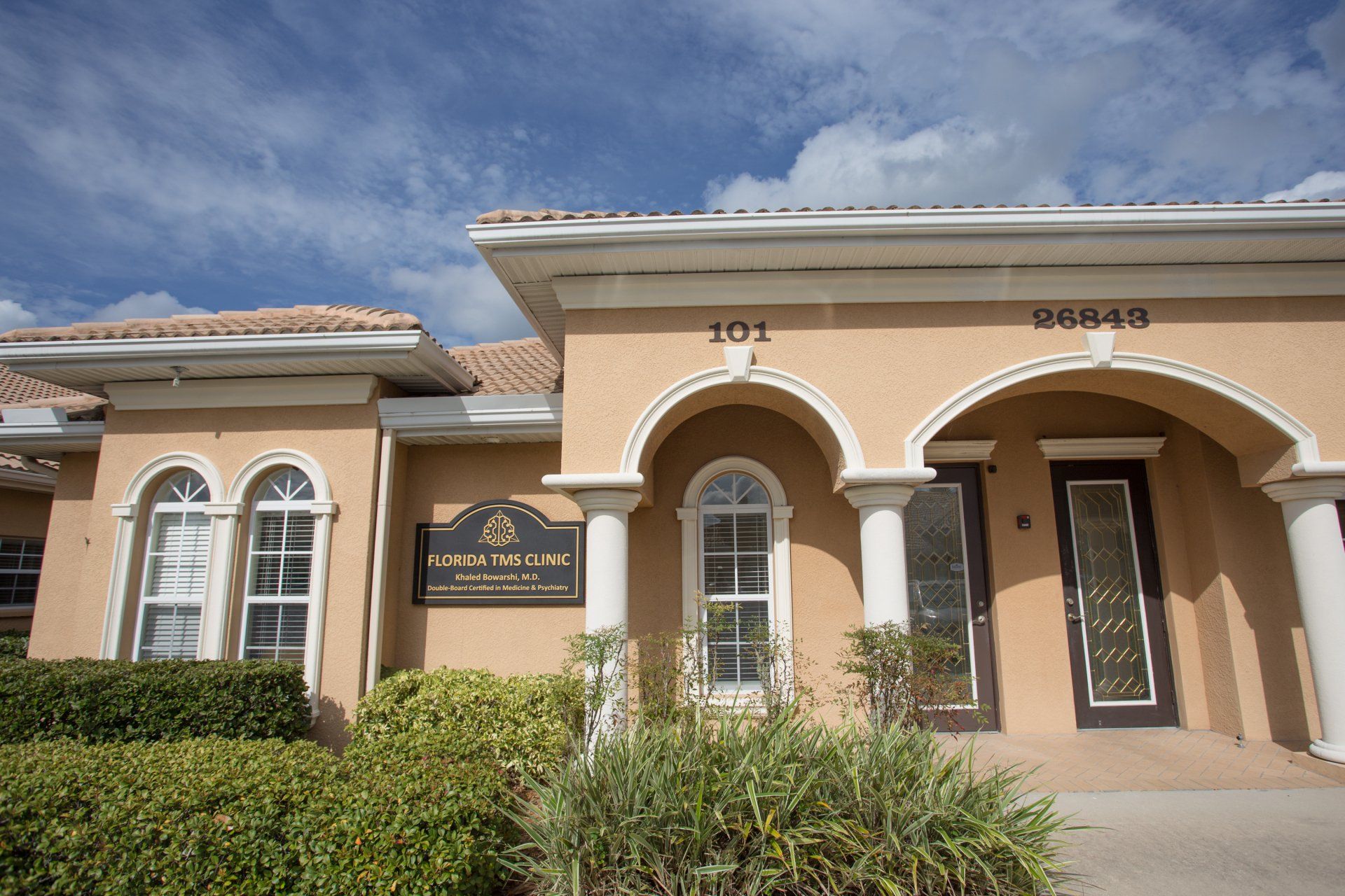 Tan building with arched entryways, address 101, and sign, under a partly cloudy blue sky. Bushes and plants in front.