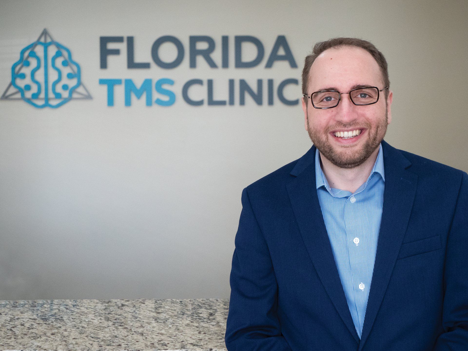 A man in a blue suit and glasses smiles, standing in front of the Florida TMS Clinic logo.
