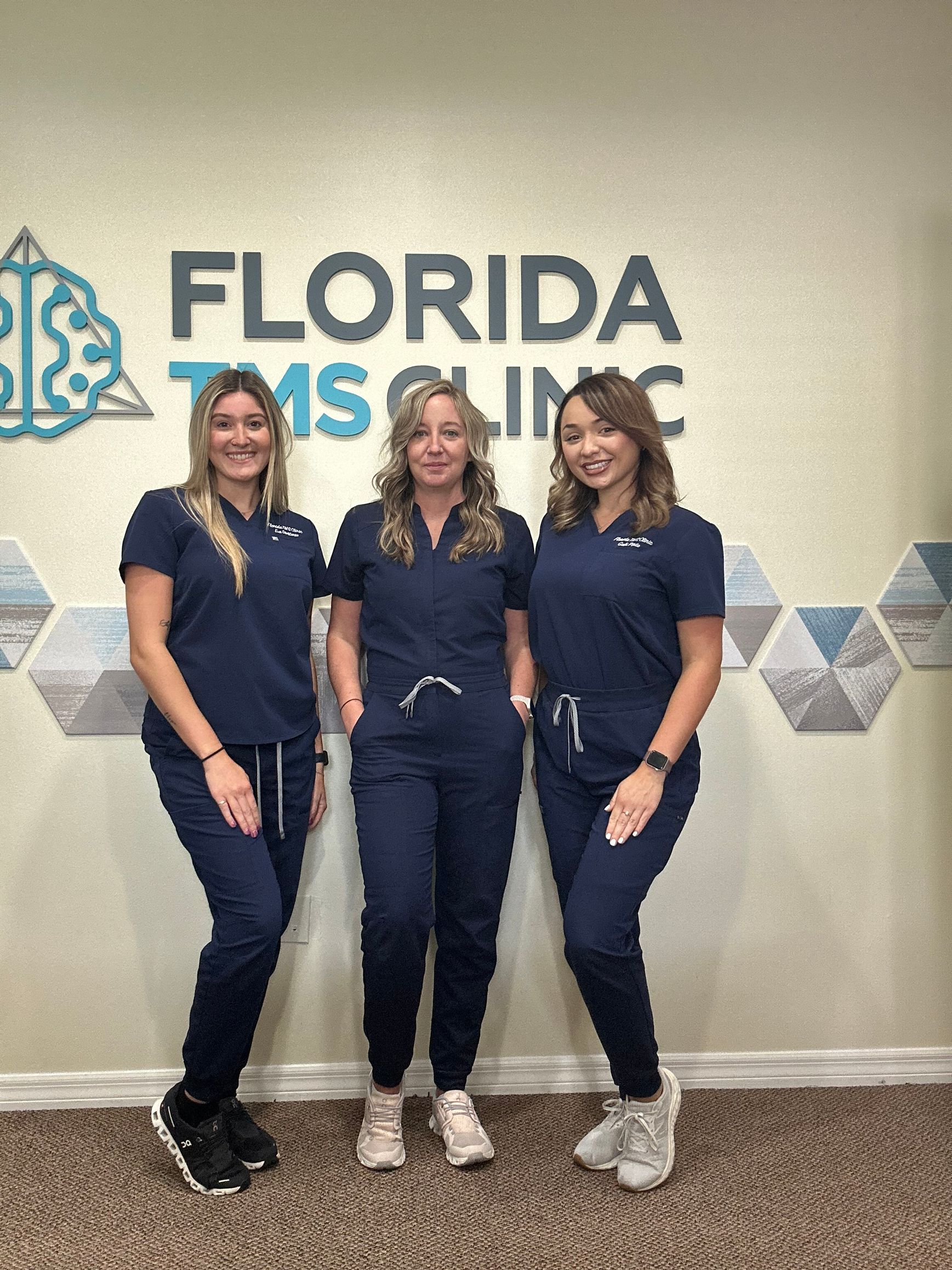 Three women in blue scrubs stand in front of the Florida TMS Clinic sign.
