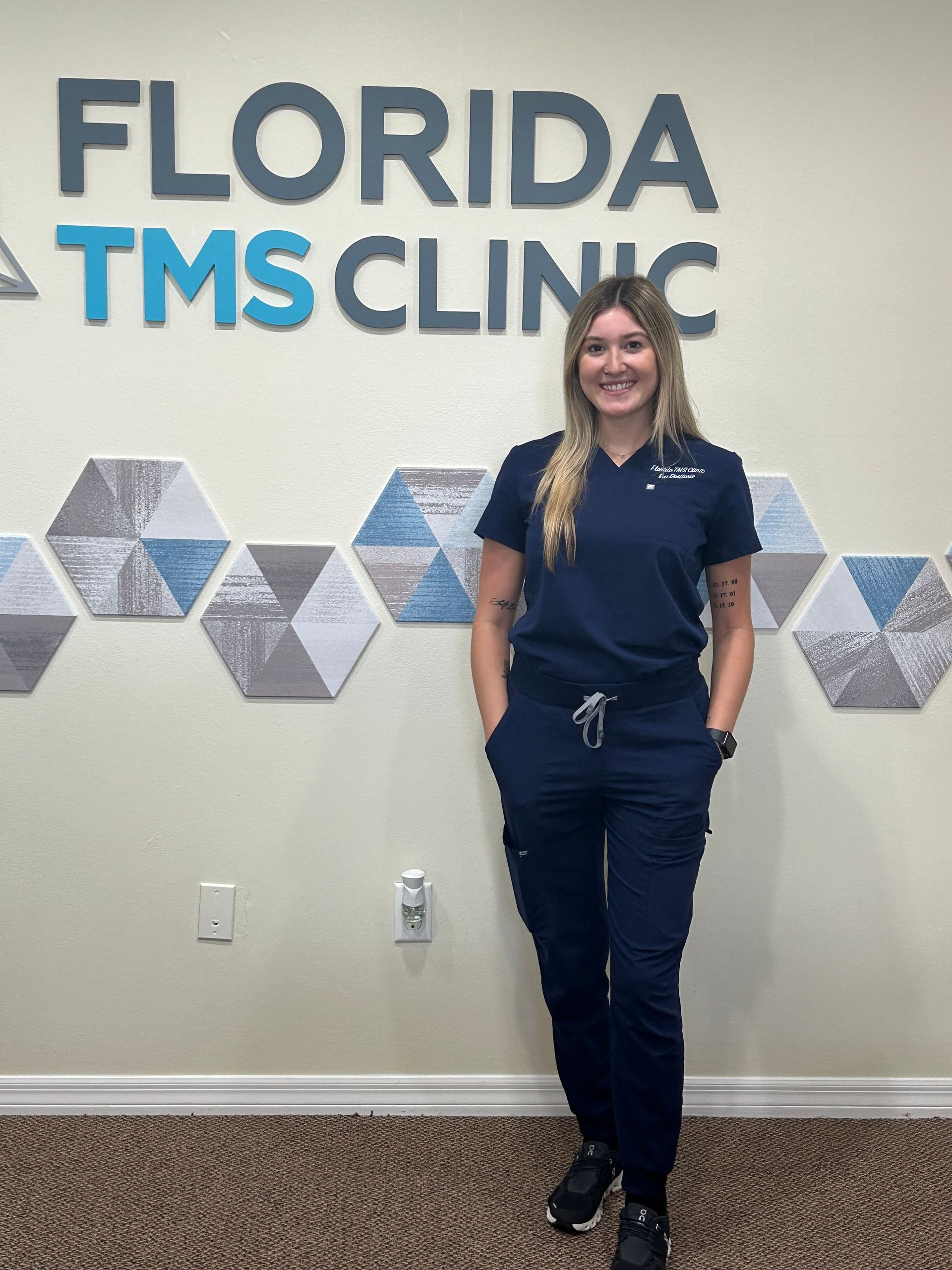 Woman in navy scrubs stands in front of the Florida TMS Clinic sign.