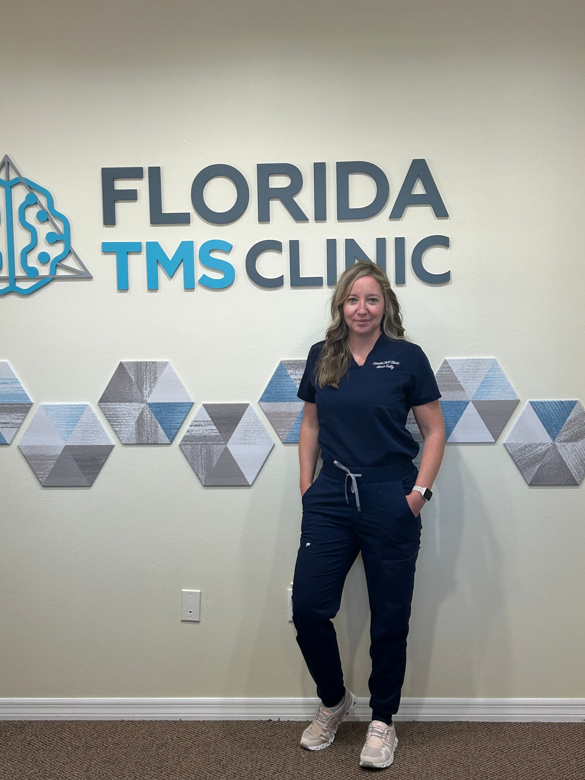 Woman in scrubs stands in front of a wall with the Florida TMS Clinic logo.