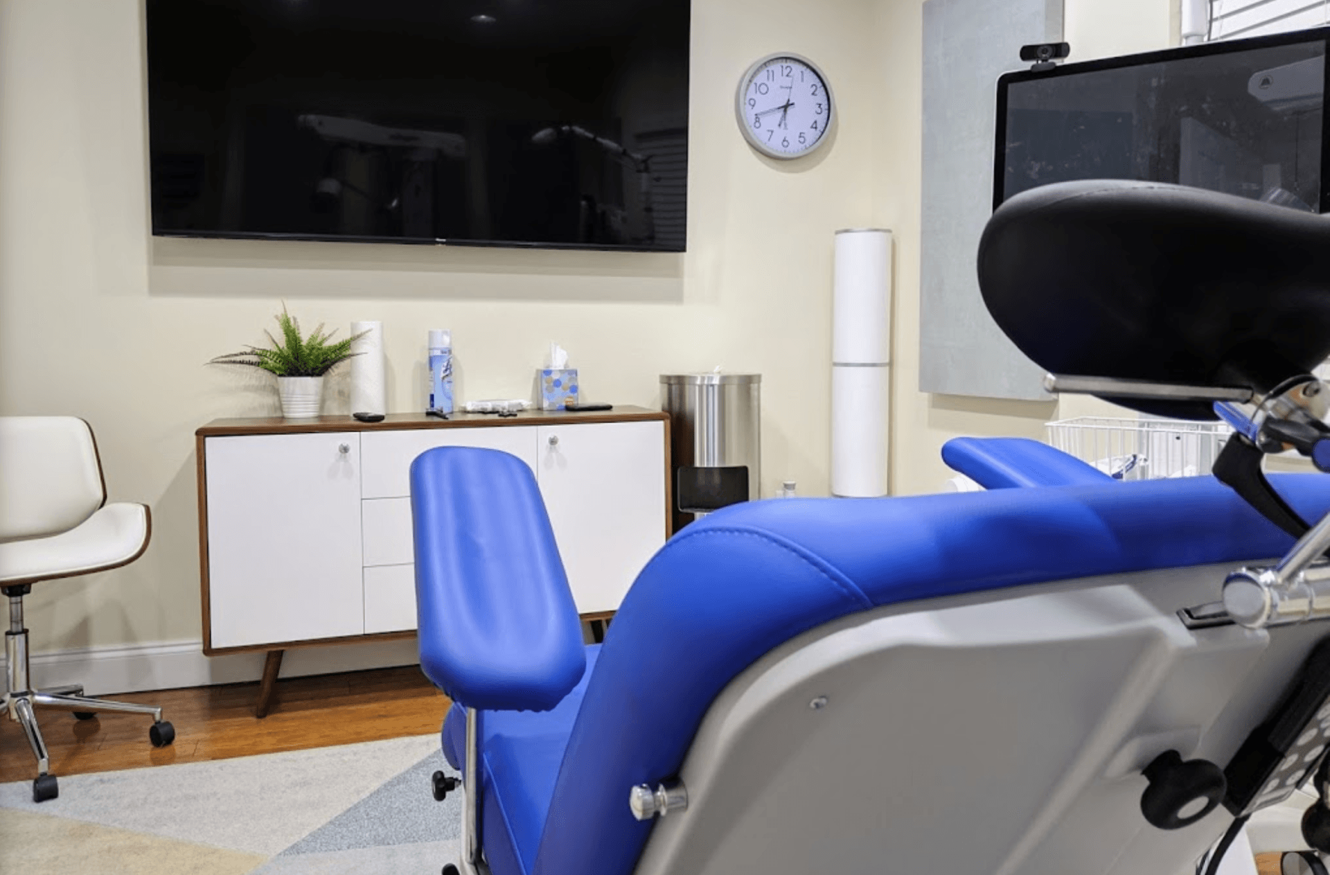 A medical examination room with a blue chair, a TV, and a white cabinet. A clock and plant are mounted on the wall.