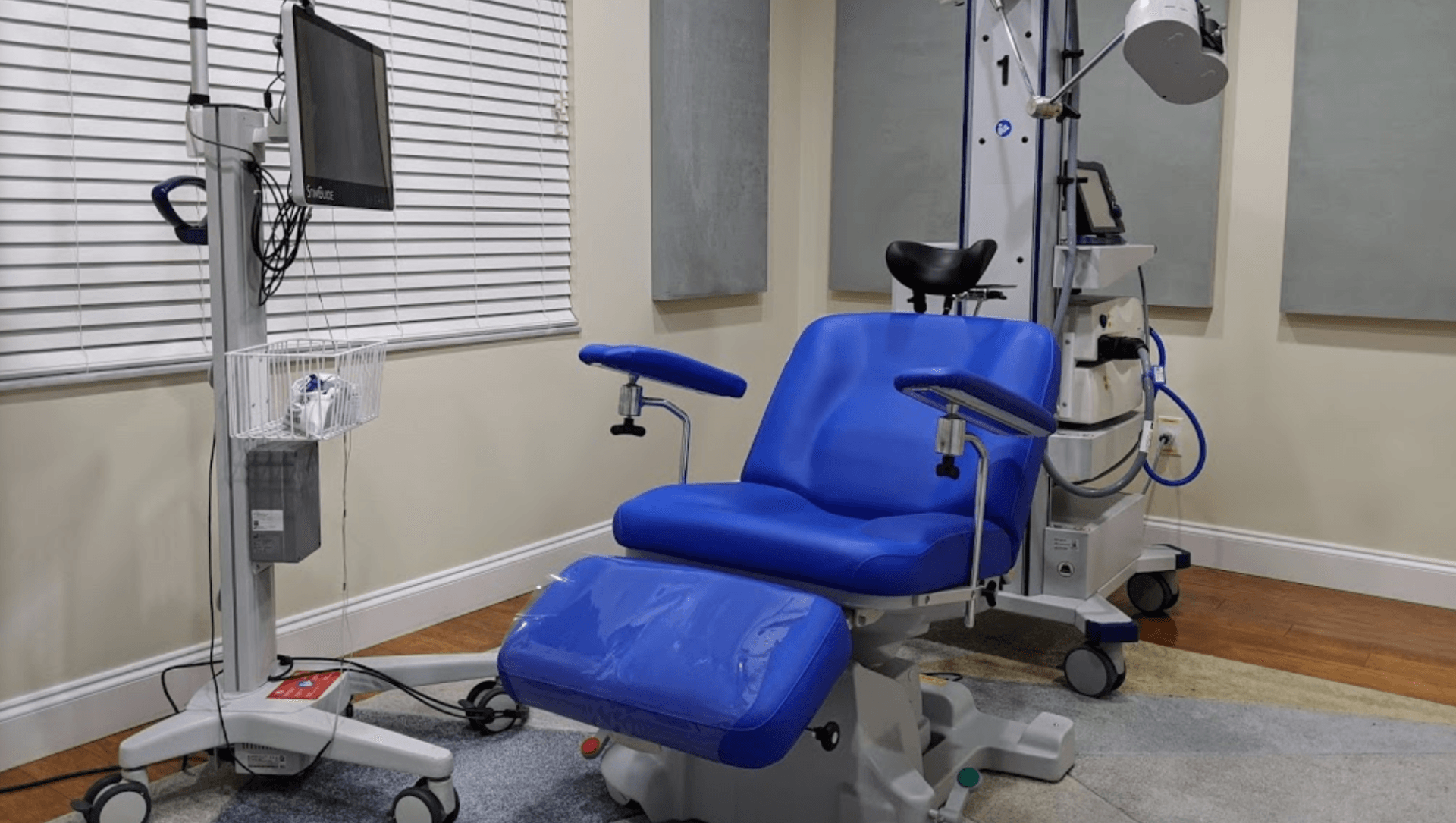 Blue examination chair with attached medical equipment; in a doctor's office, with a monitor and rolling cart.