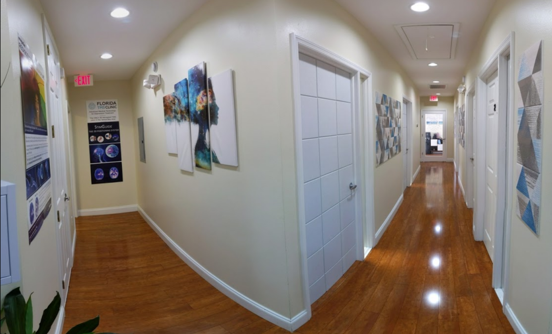 A bright hallway with wooden floors and off-white walls. Artwork and doors line the sides. Recessed lights illuminate the space.