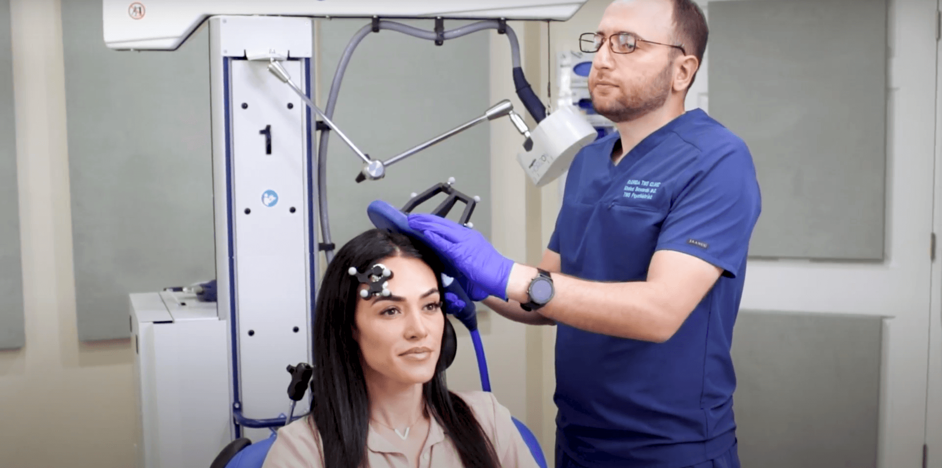 A doctor in blue scrubs adjusts a device on a woman's head. The woman sits in a chair, and the setting appears to be a medical office.