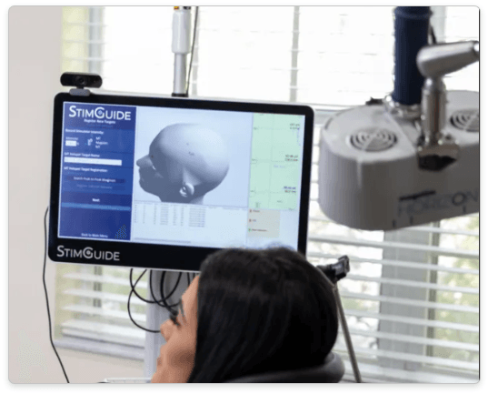 Woman facing a monitor displaying a 3D rendering of a head. A StimGuide system with a camera and medical equipment are visible.