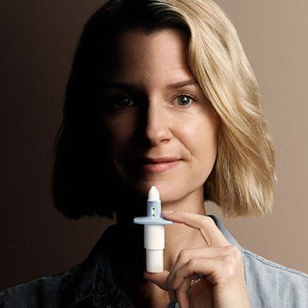 Woman holding a nasal spray, smiling, against a neutral background.