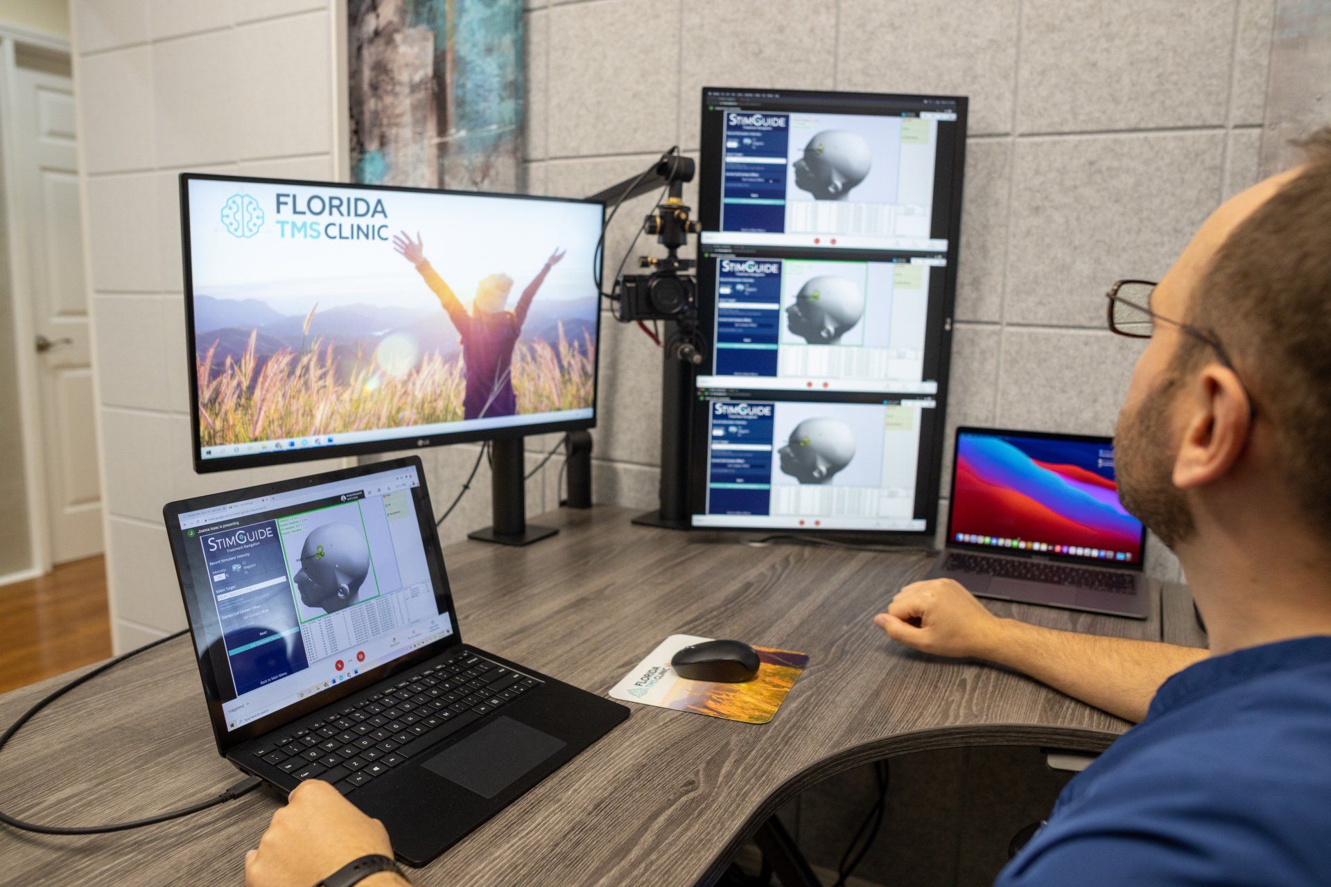 A man with glasses works at a desk with multiple monitors displaying 3D medical images. The setup includes a laptop and a larger monitor with the Florida Eye Clinic logo.