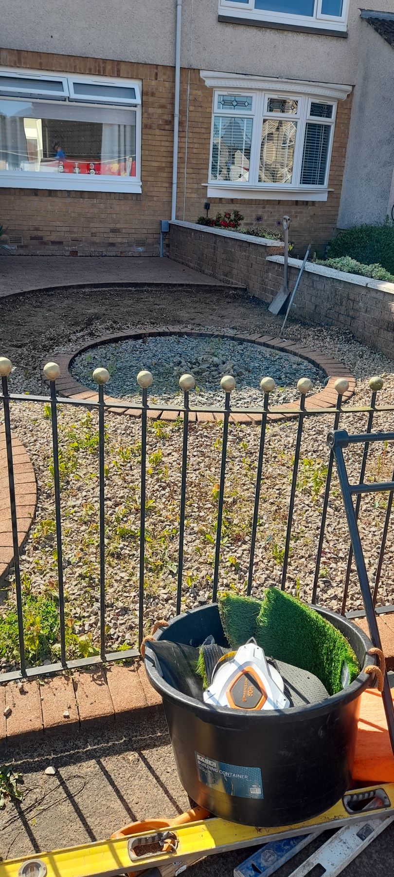 Front yard with black metal fence, leaf-covered soil bed, and a black bucket of gardening debris on a patio