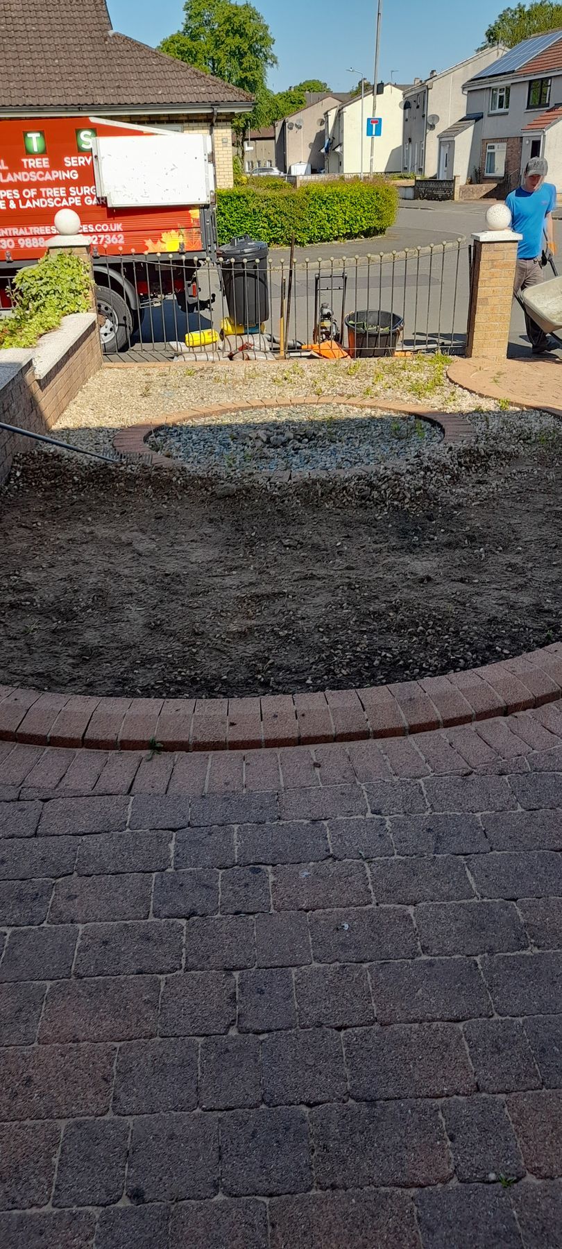 Small landscaped roundabout with flowers, hedge, and brick paving in a sunny residential street