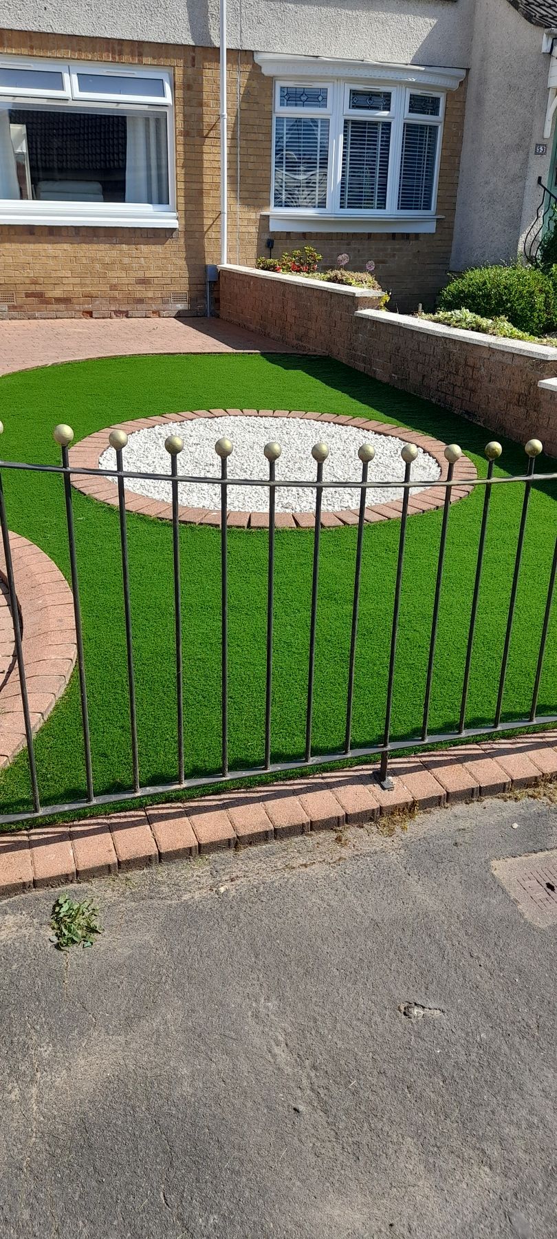Front garden with bright green lawn, circular gravel bed, brick edging, and a low black metal fence by a house.