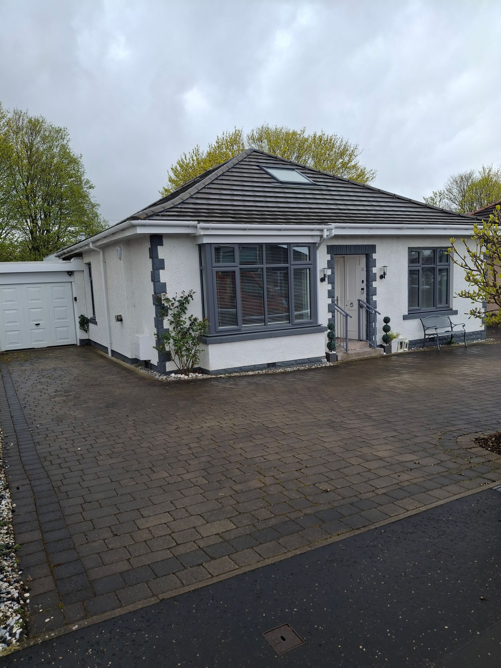 Single-story white house with a gray roof, front windows, and a paved driveway on a cloudy day