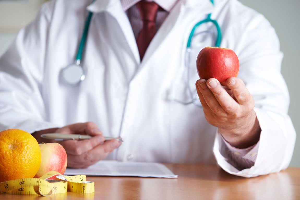 Il medico in camice bianco tiene in mano una mela rossa, sulla scrivania ci sono frutta e un metro a nastro.