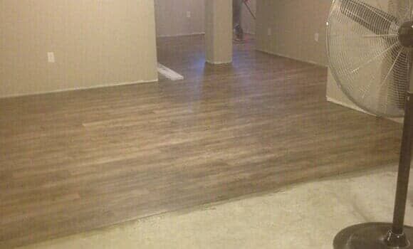 A room transition with wood-look laminate flooring adjacent to light-colored carpet, viewed near a pedestal fan.