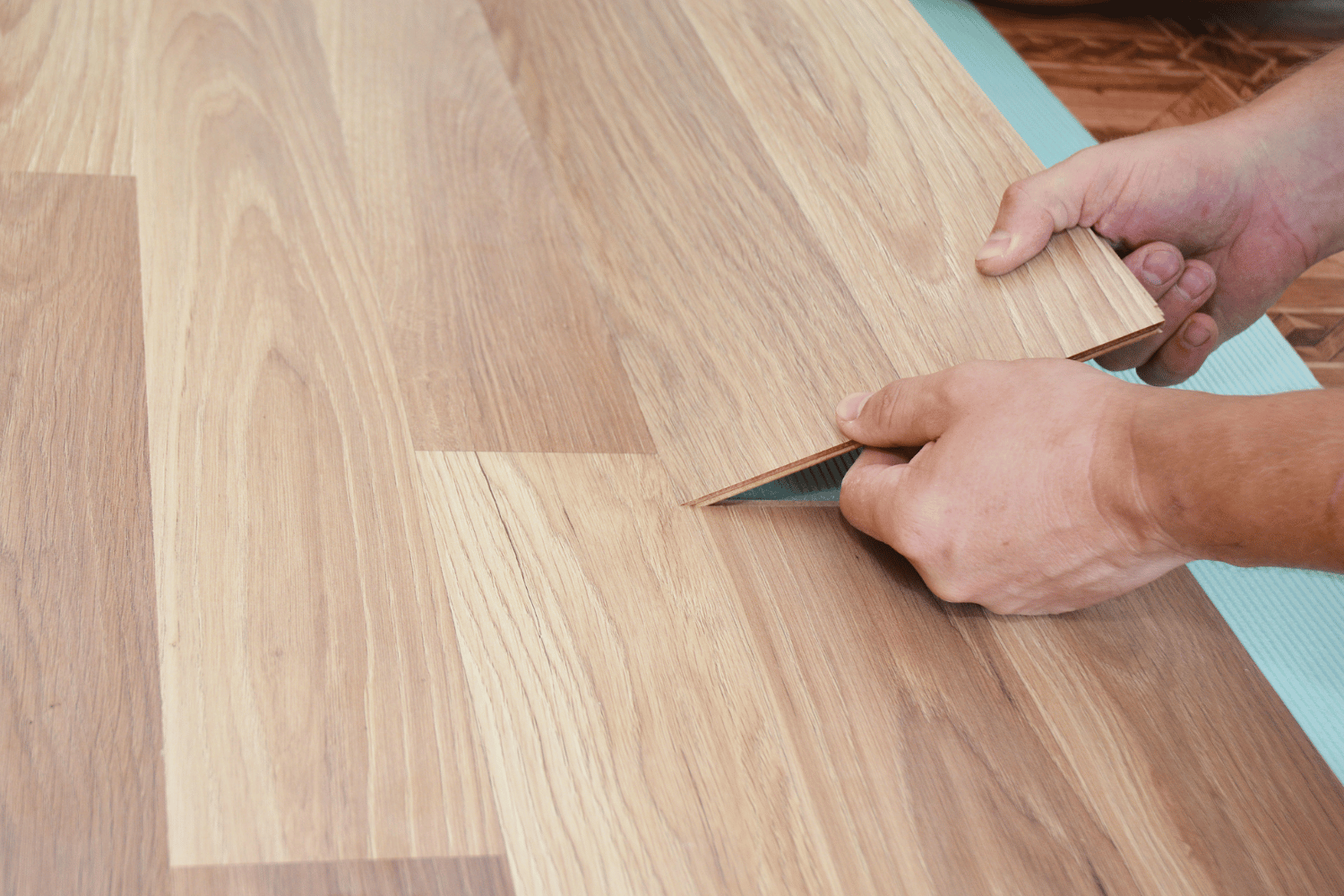 Hands holding and positioning a wood-look laminate flooring plank onto an underlayment layer during installation.
