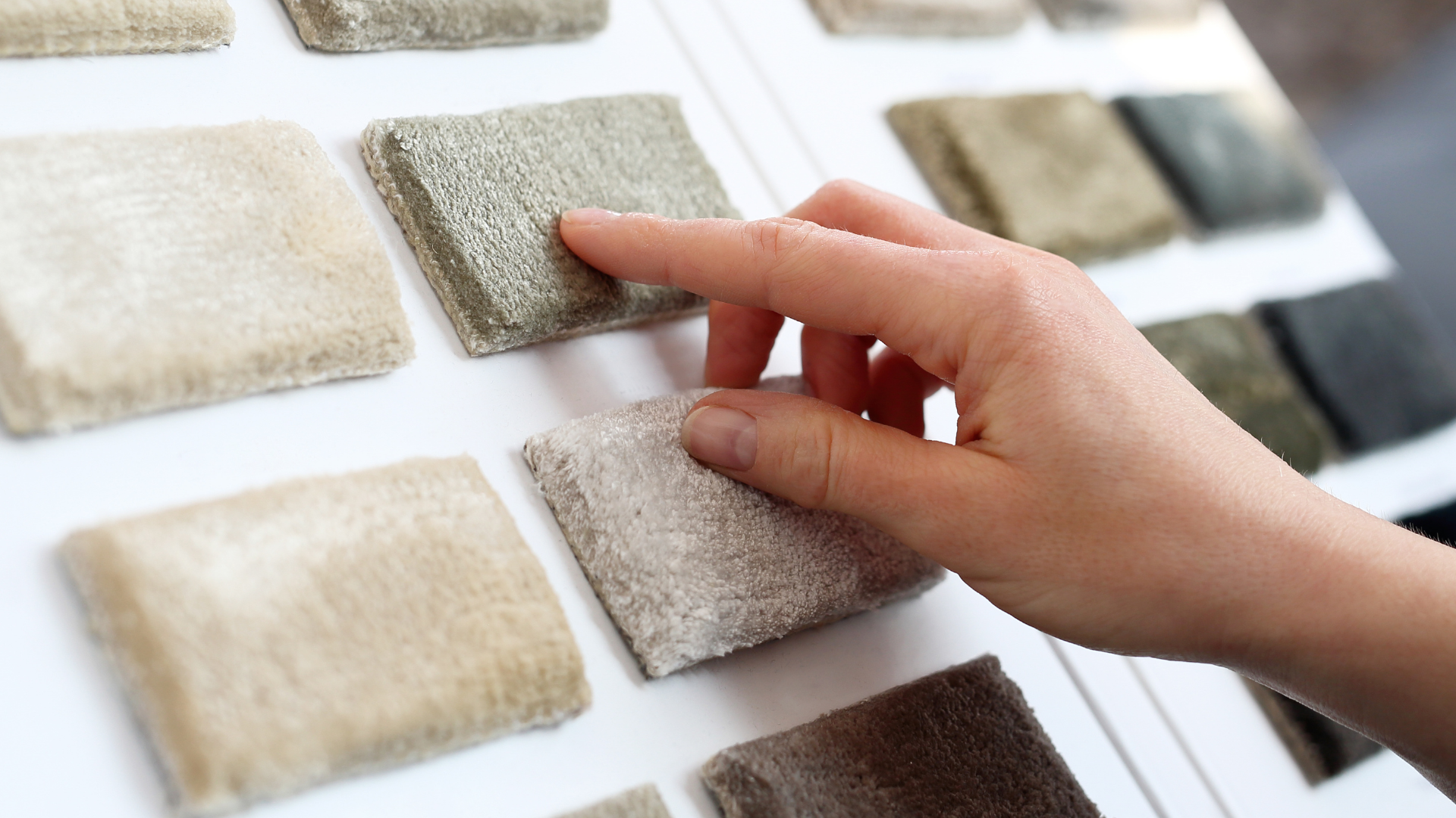 A hand touches a sample from a display of various square, textured carpet swatches in neutral tones.