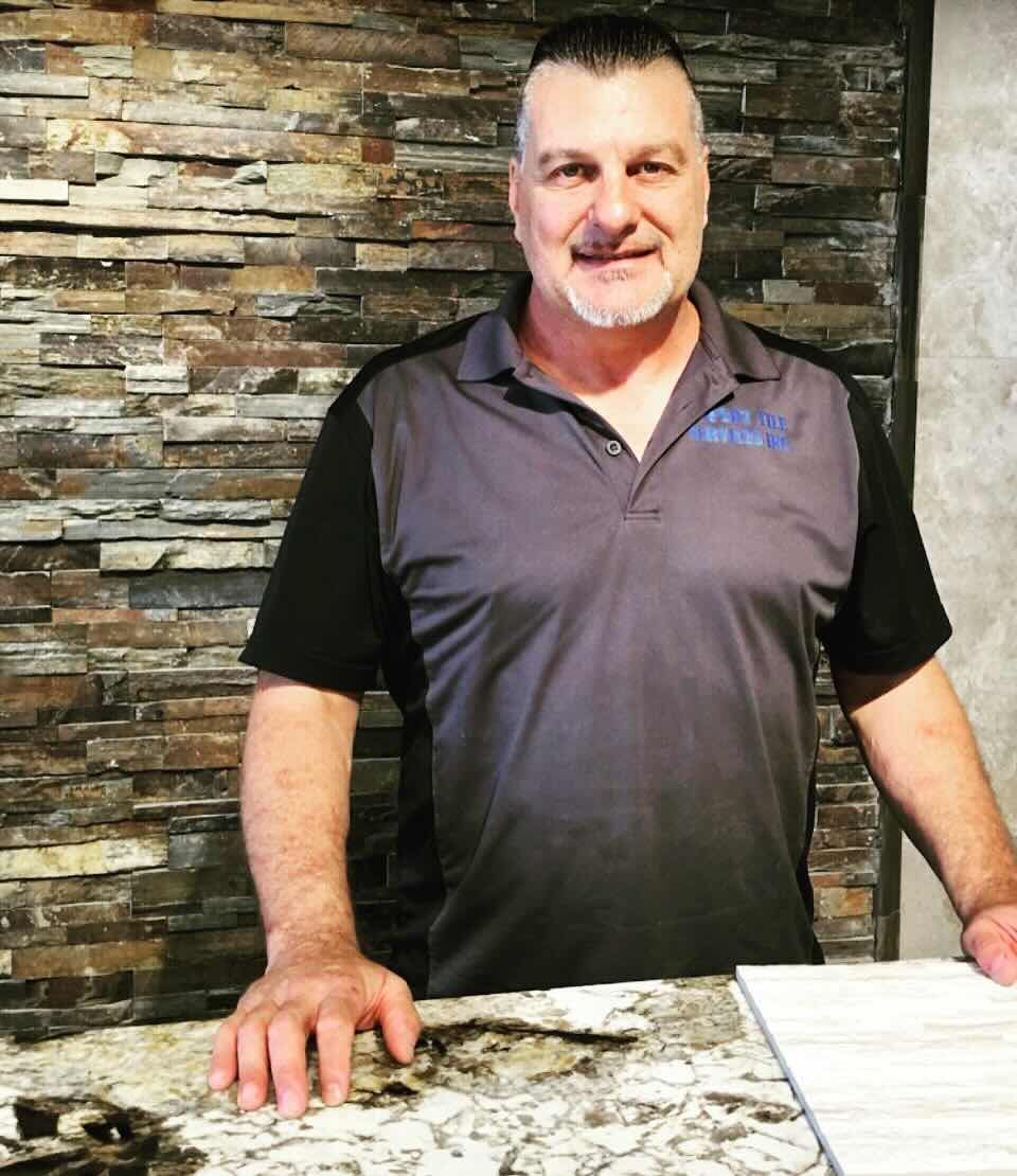 A man in a gray shirt is standing in front of a stone wall.