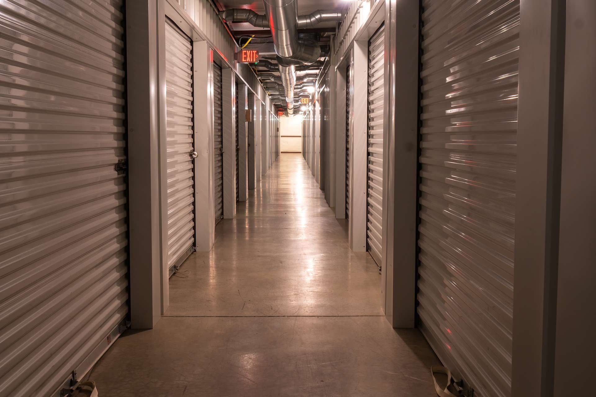 Interior climate controlled rental storage units with exit signs and locked metal roller doors. Hallway with rows of storage unit doors on both sides. Concrete floor, overhead pipes, and dim lighting.