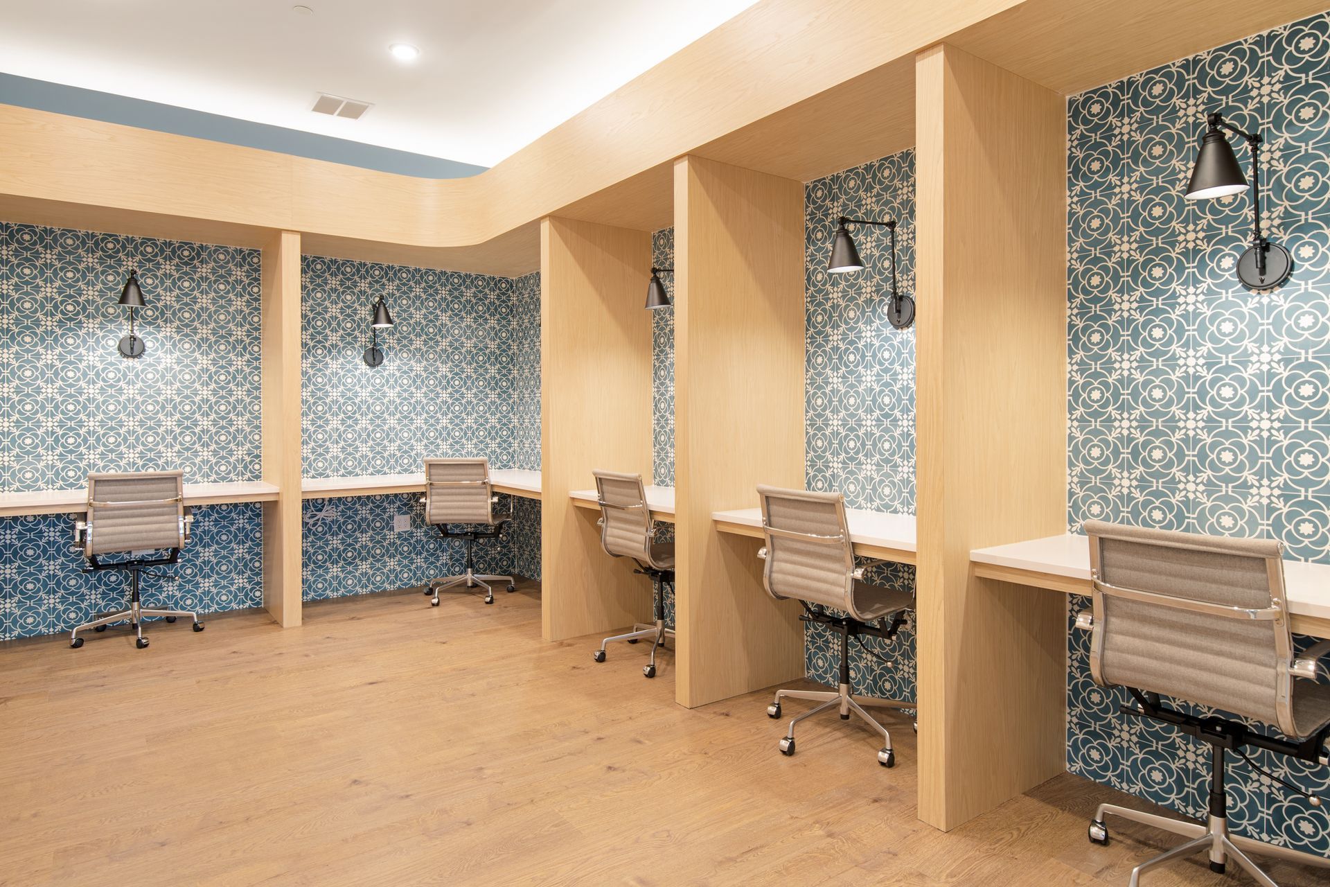 Work cubicles with teal patterned walls, light wood desks, and gray office chairs, lit by black wall lamps.