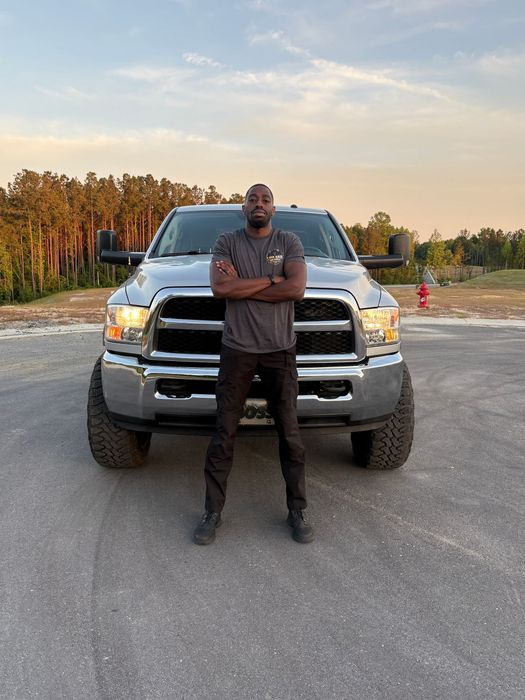 Man standing in front of a silver pickup truck on a gravel road at sunset