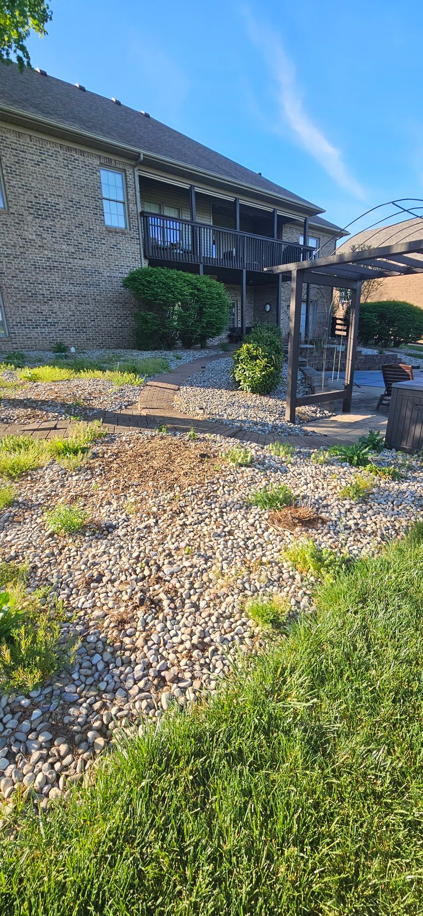 Stone house with balcony, gravel yard, and green lawn under a blue sky.
