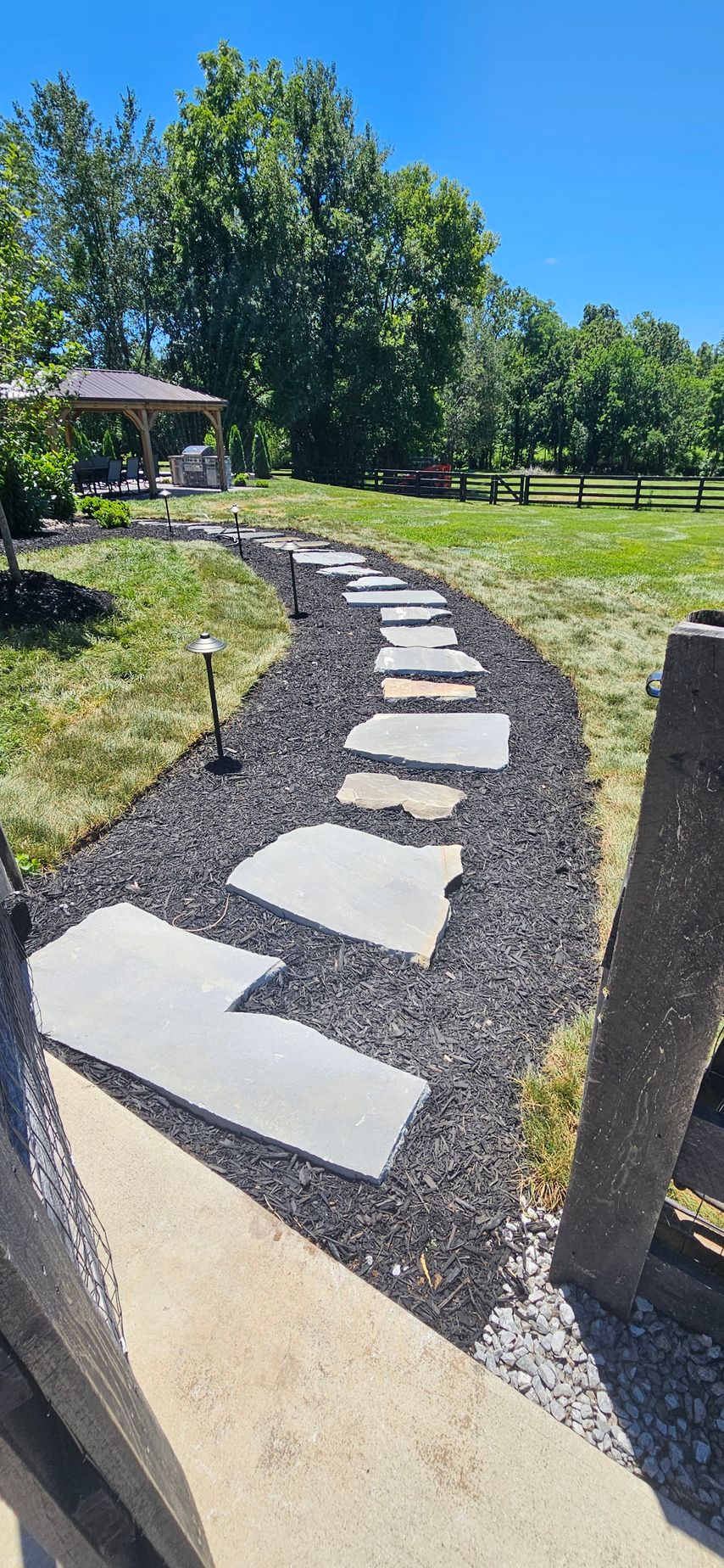 Curved stone stepping path through a grassy park under blue sky, with trees and a bench nearby