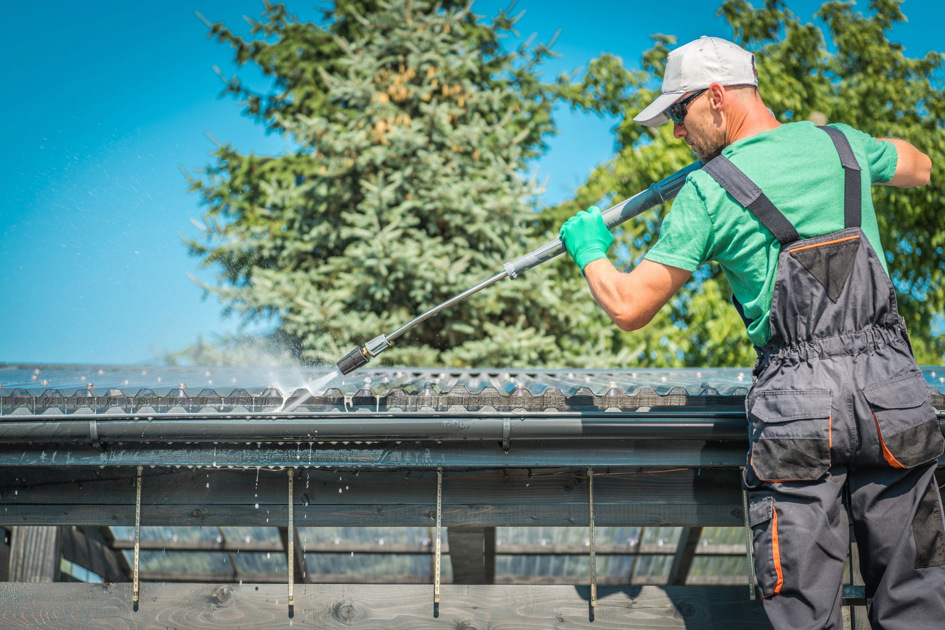 A person in work clothes and a cap uses a pressure washer to clean a corrugated roof outdoors on a sunny day.