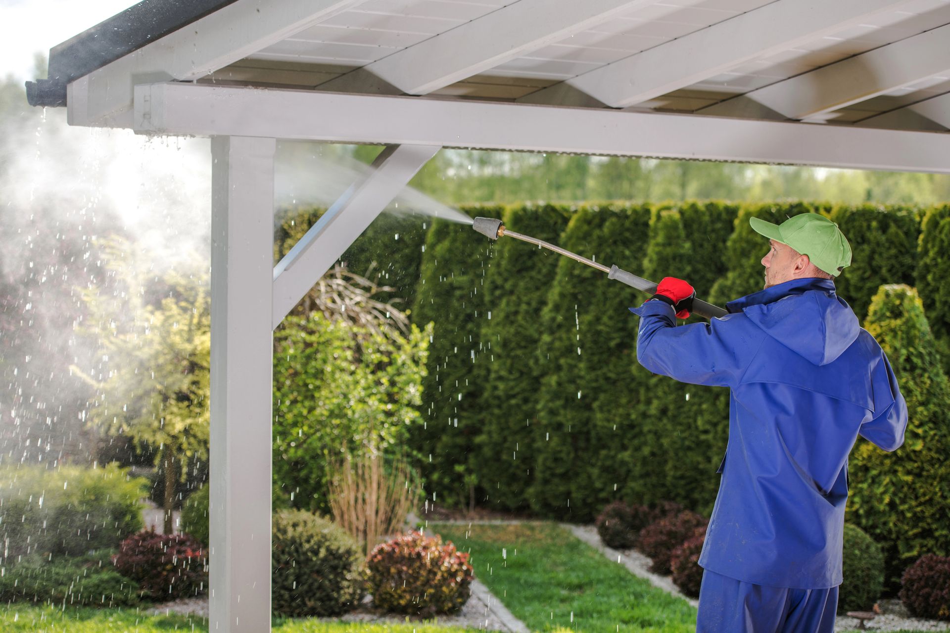 A person wearing a blue rain suit and green cap power washes a white wooden pergola in a backyard garden.