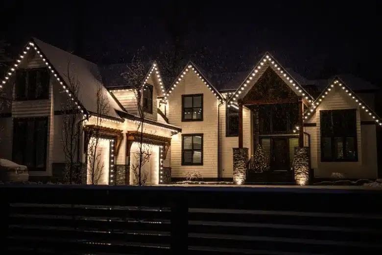 House outlined with warm white string lights at night, viewed from across a dark fence.