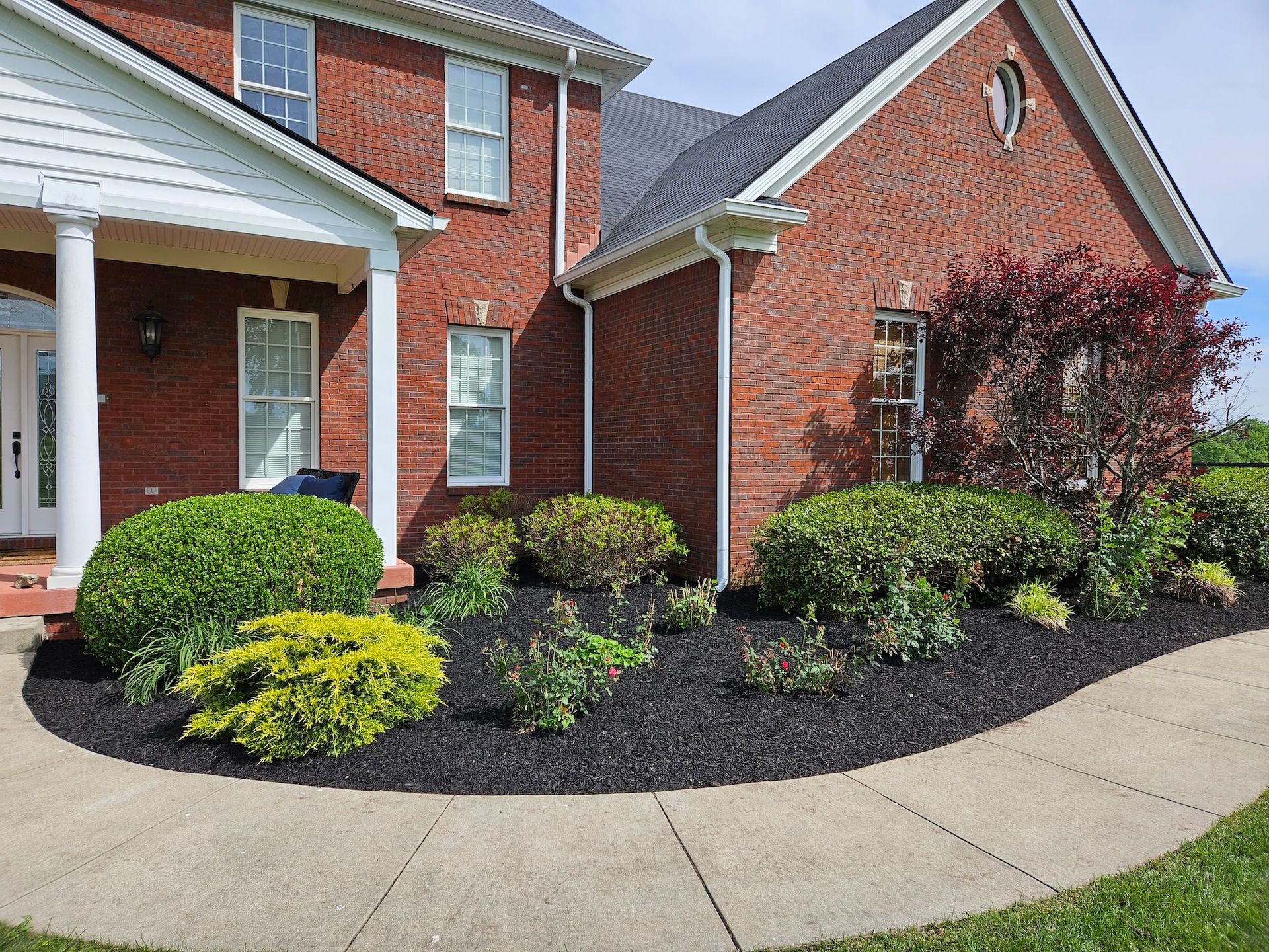 Red brick house with a curved walkway and landscaped garden beds filled with greenery and black mulch.