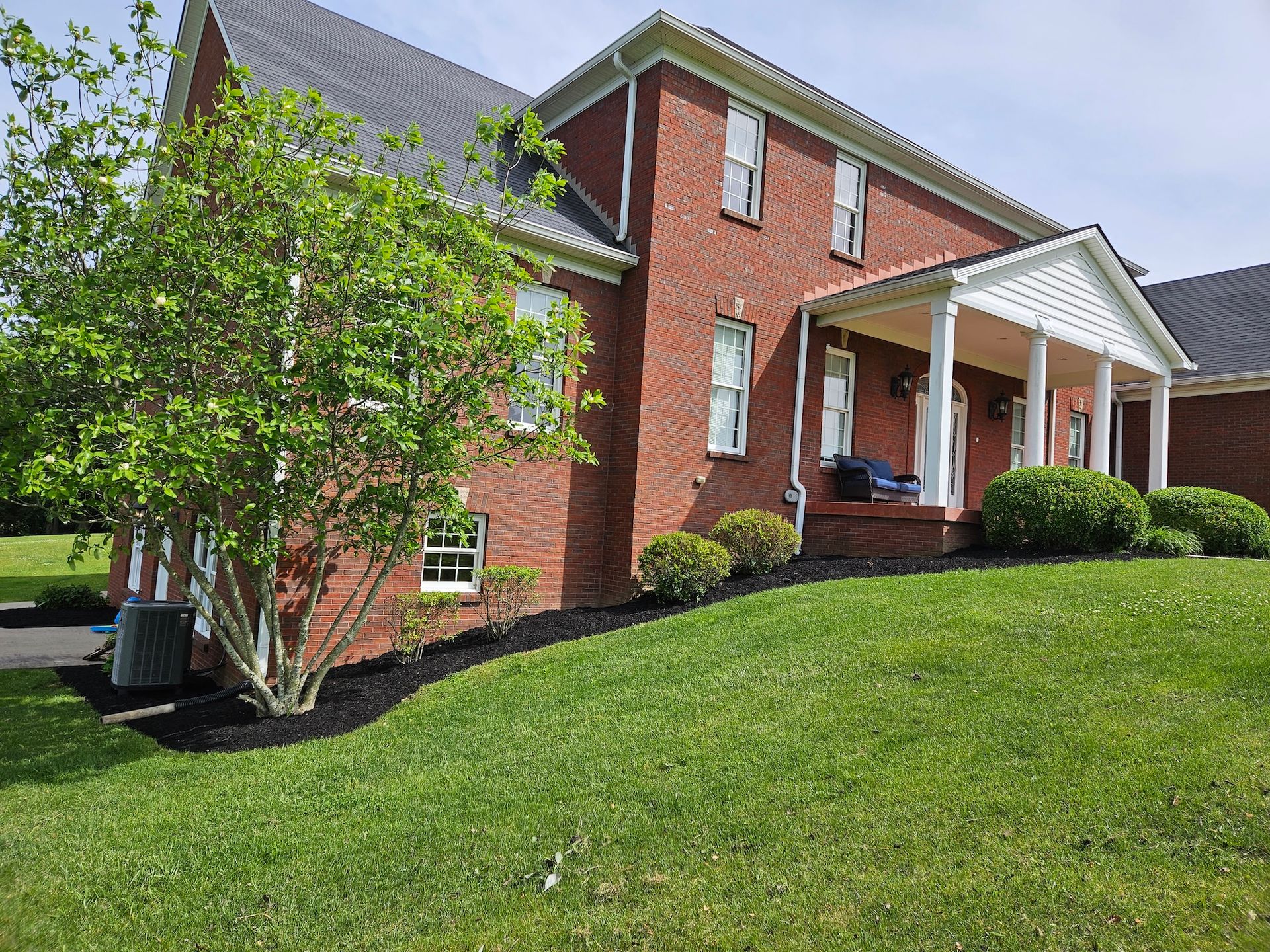 Two-story brick house with a white porch and pillars. Lush green lawn with black mulch border.