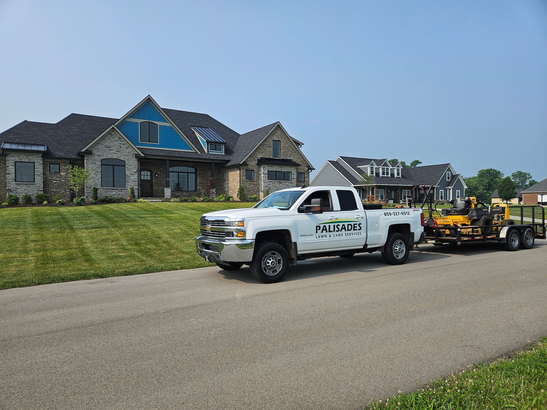 White pickup truck with trailer and lawn equipment parked in front of a house on a sunny day.