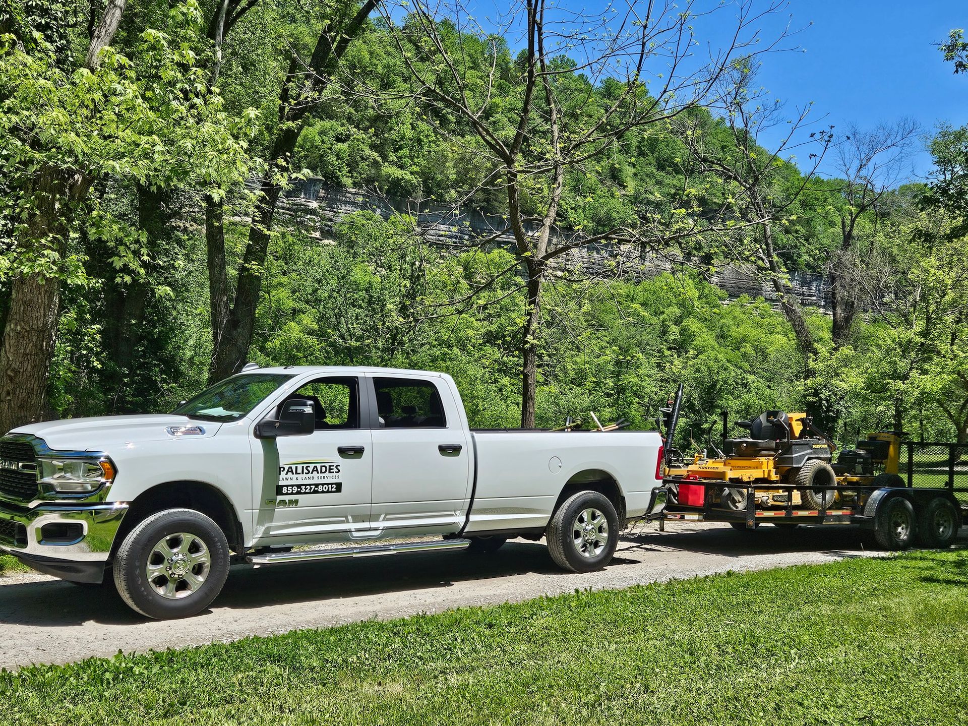White pickup truck towing equipment trailer on a gravel road, trees and cliff in background.