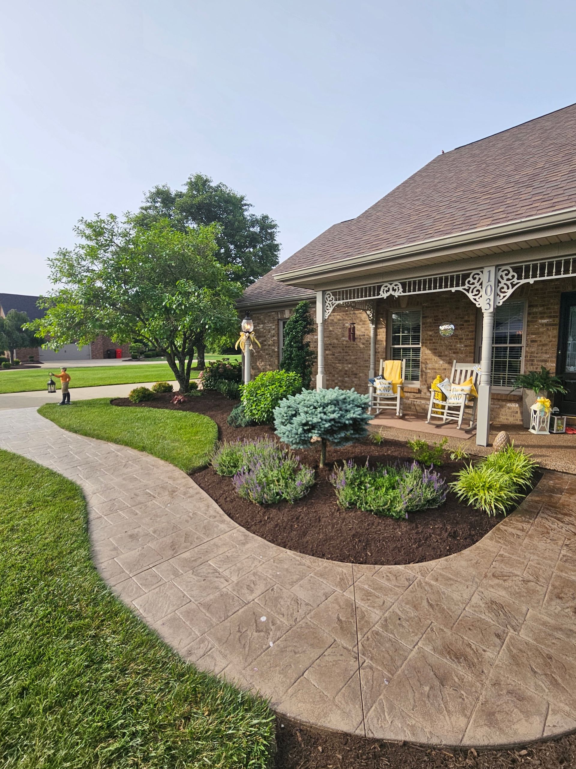 Stone walkway curves towards a brick house with a porch and landscaped flower beds.