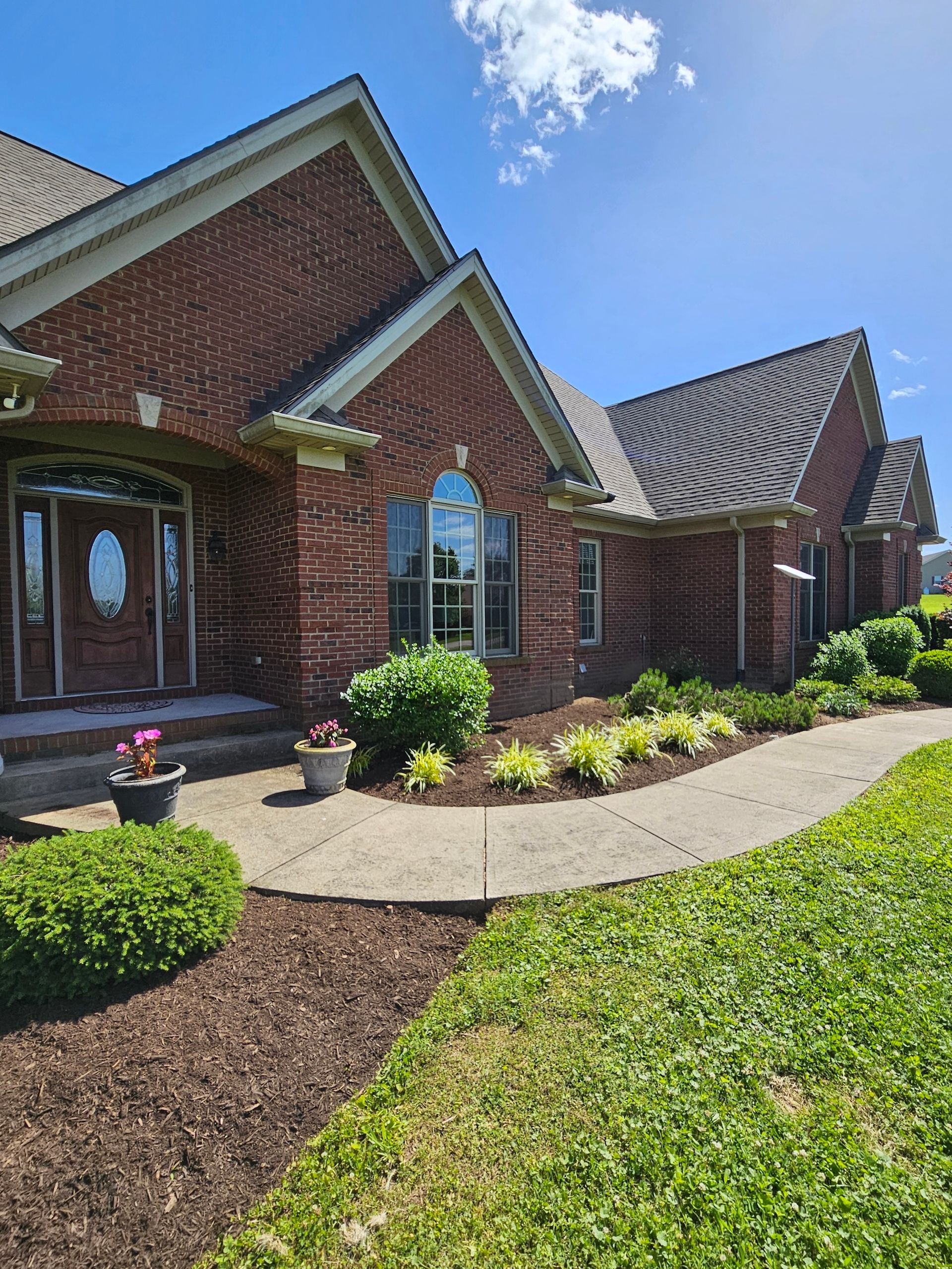Red brick house with a curved walkway, surrounded by landscaping, under a sunny sky.