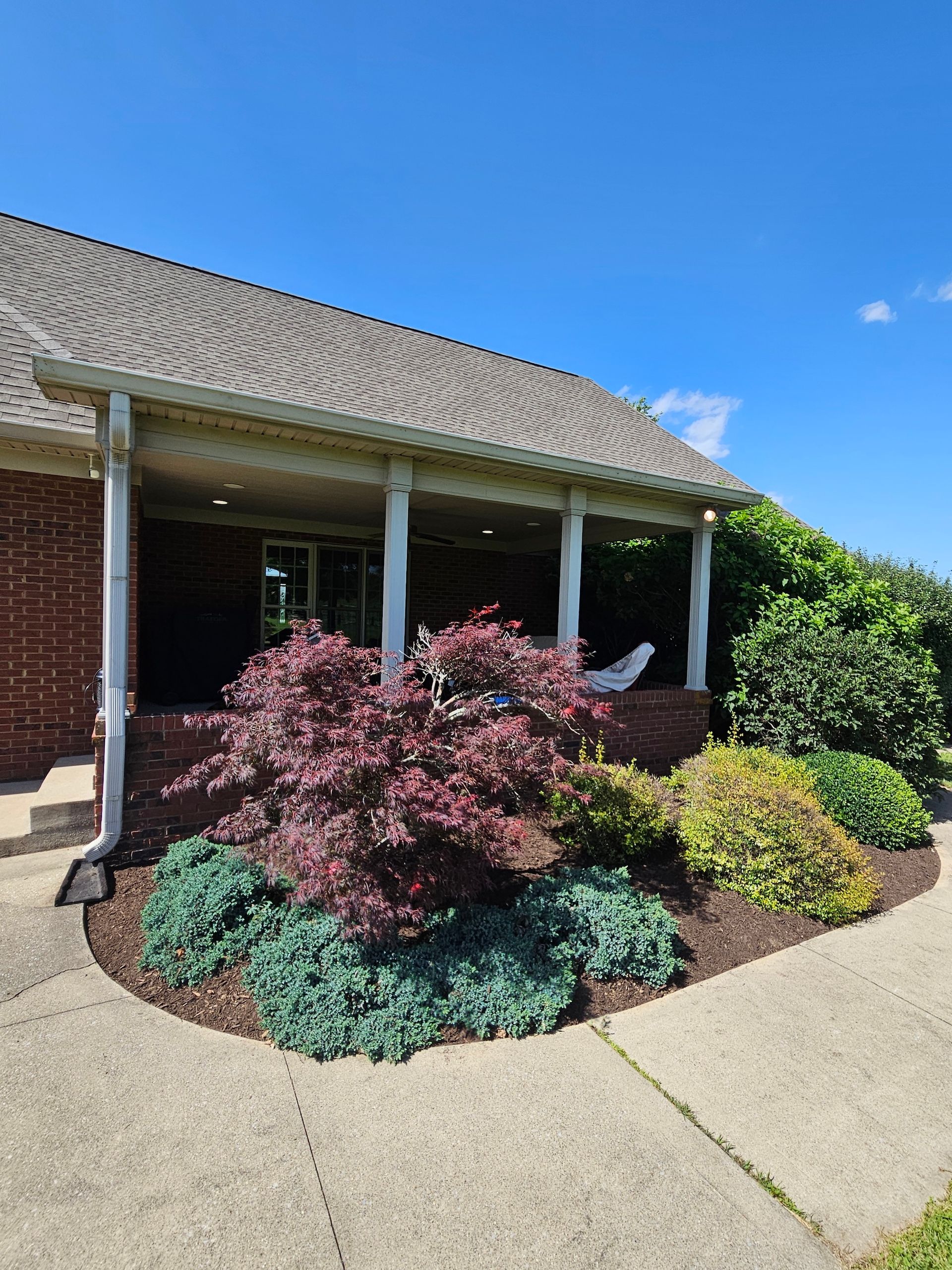 A house with a brick facade and a covered porch, featuring a Japanese maple and other shrubs in a landscaped bed.