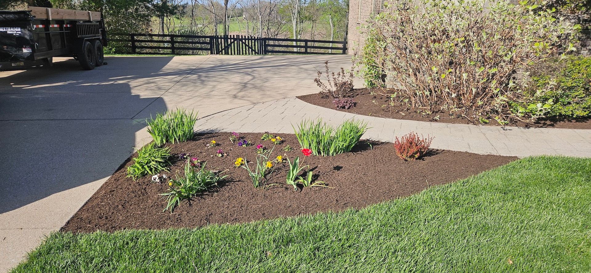 A flower bed with mulch, small colorful flowers, and green grass next to a paved driveway.