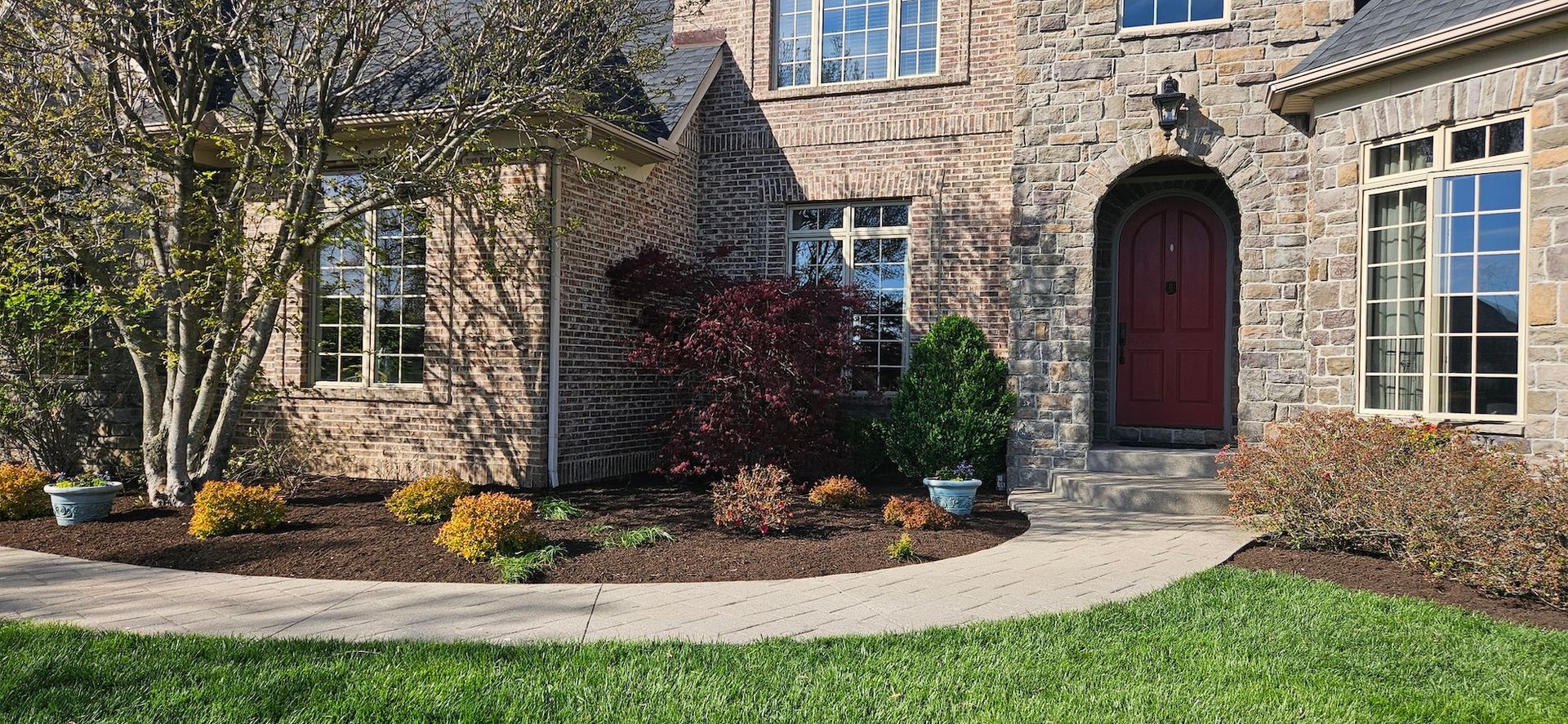 Stone house with arched entryway and red door, surrounded by landscaping and a curving walkway.