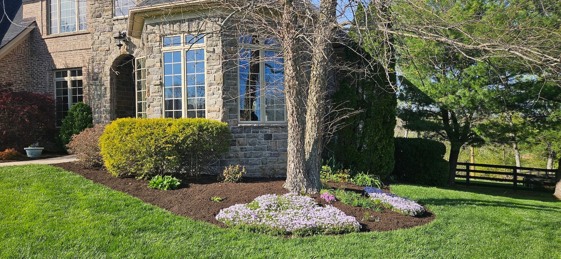 A stone house with a lush green lawn, trees, and colorful flower beds on a sunny day.