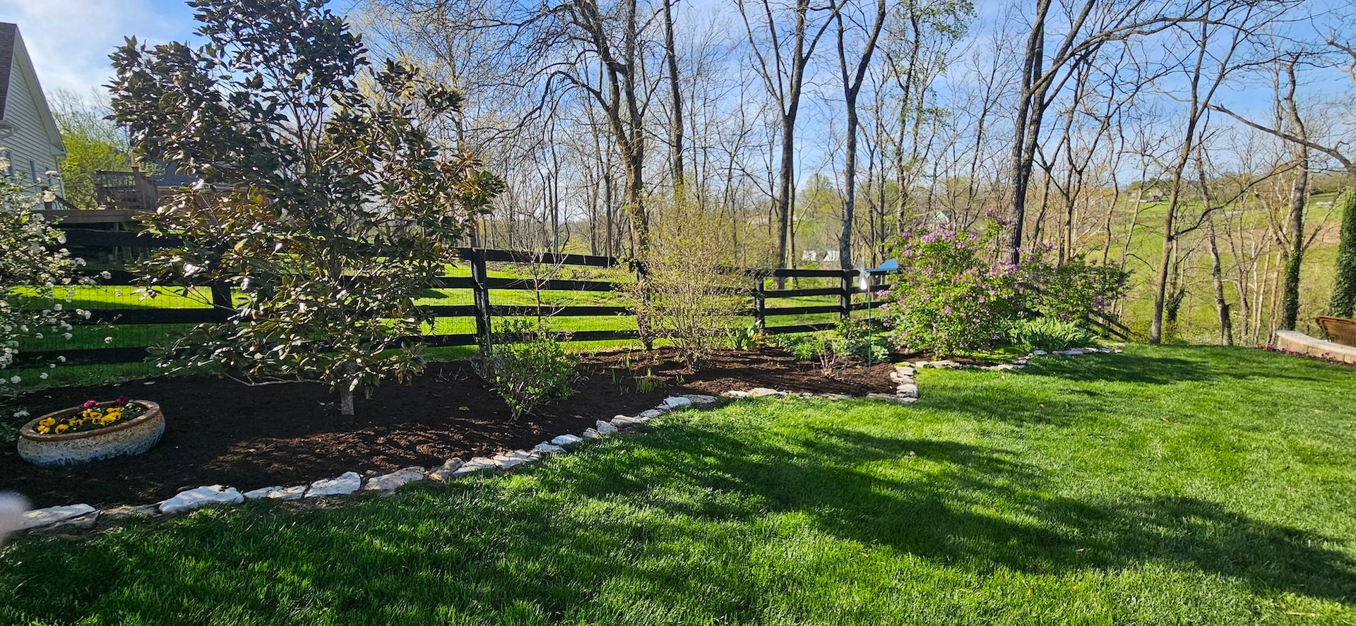 Green lawn bordered by mulch, white stones, and trees. A black fence separates the yard from a wooded area.