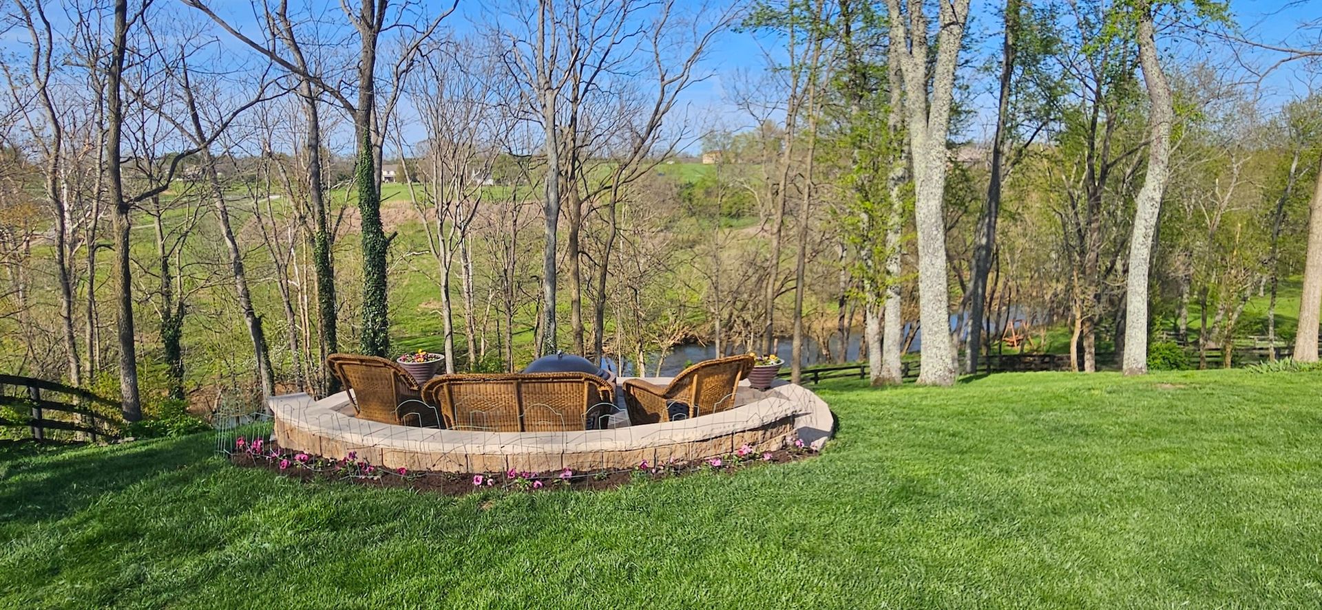 A fire pit with seating in a grassy backyard, surrounded by trees and a blue sky.