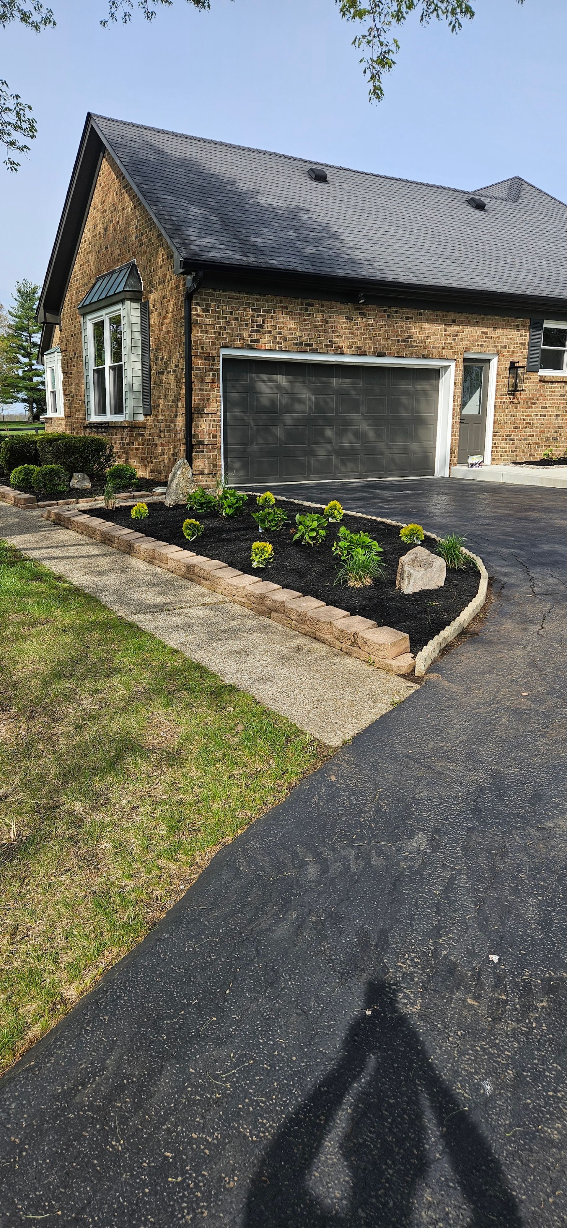 A brick house with a garage, garden bed, and a paved driveway. A shadow of a person is cast on the driveway.