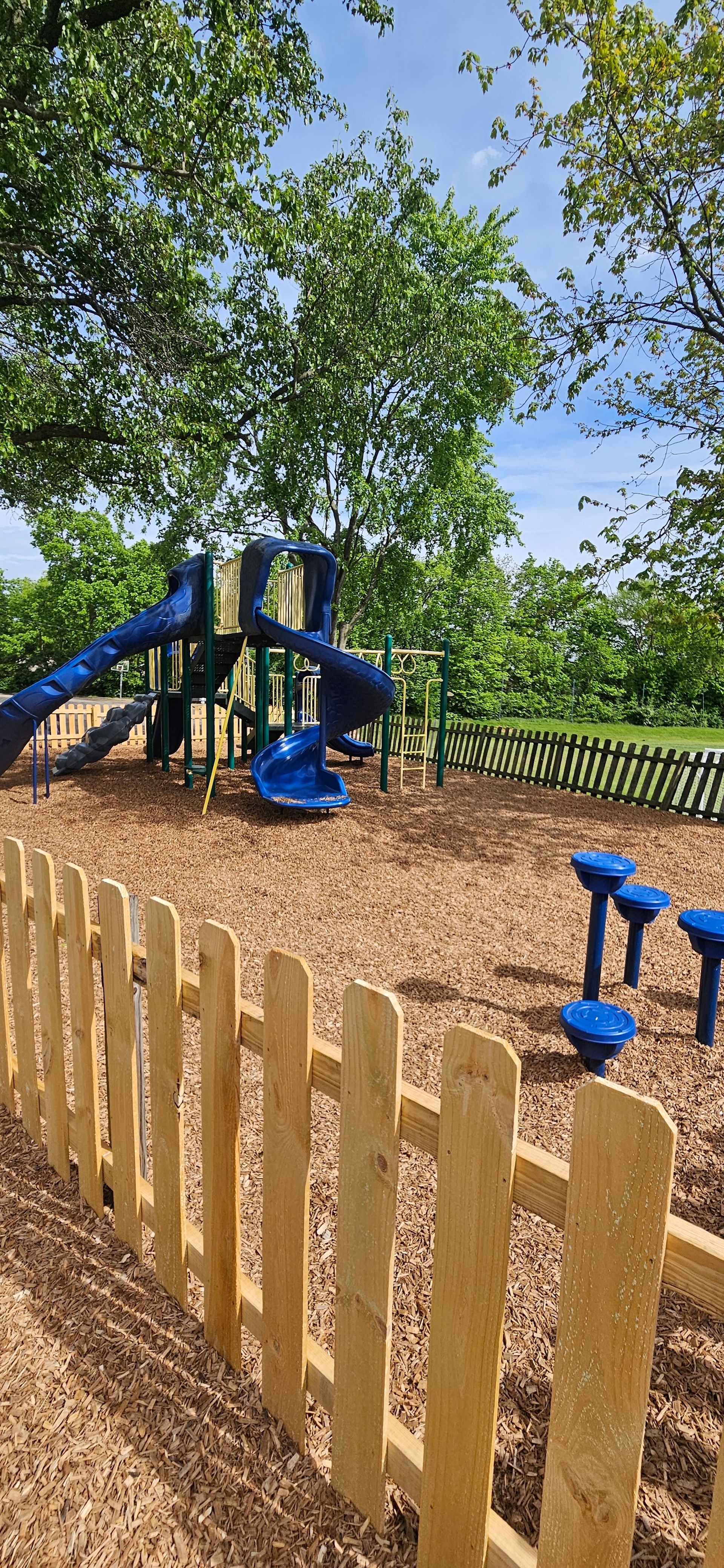 A playground with a slide, blue equipment, a wooden fence, and mulch. Trees and blue sky in the background.
