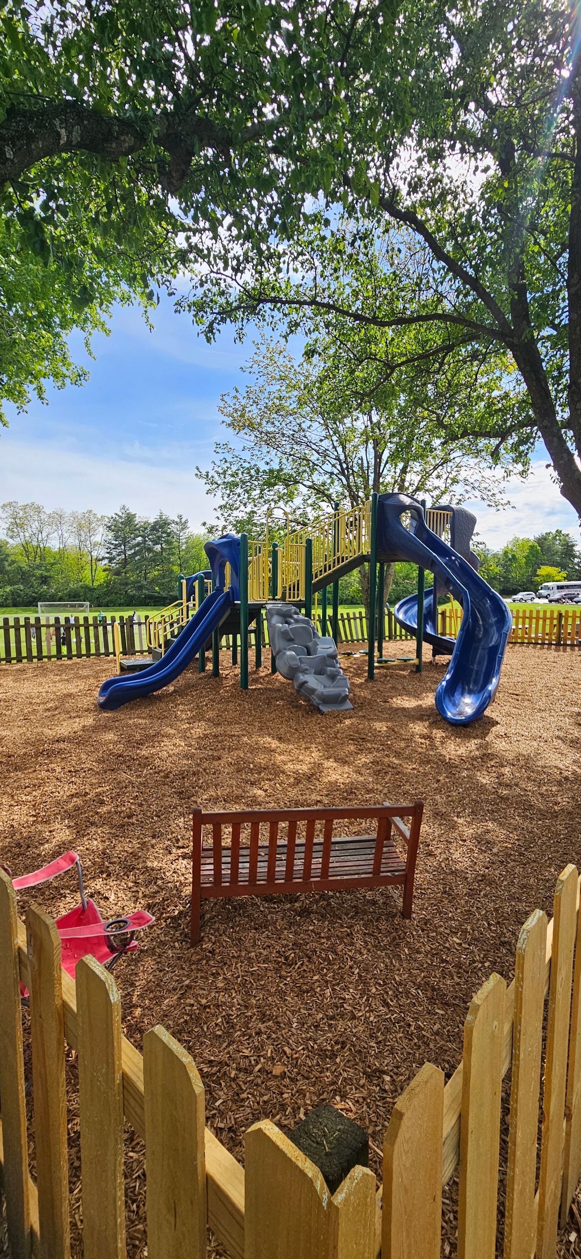 Playground with blue slides, wood chips, and a wooden fence, framed by trees under a blue sky.