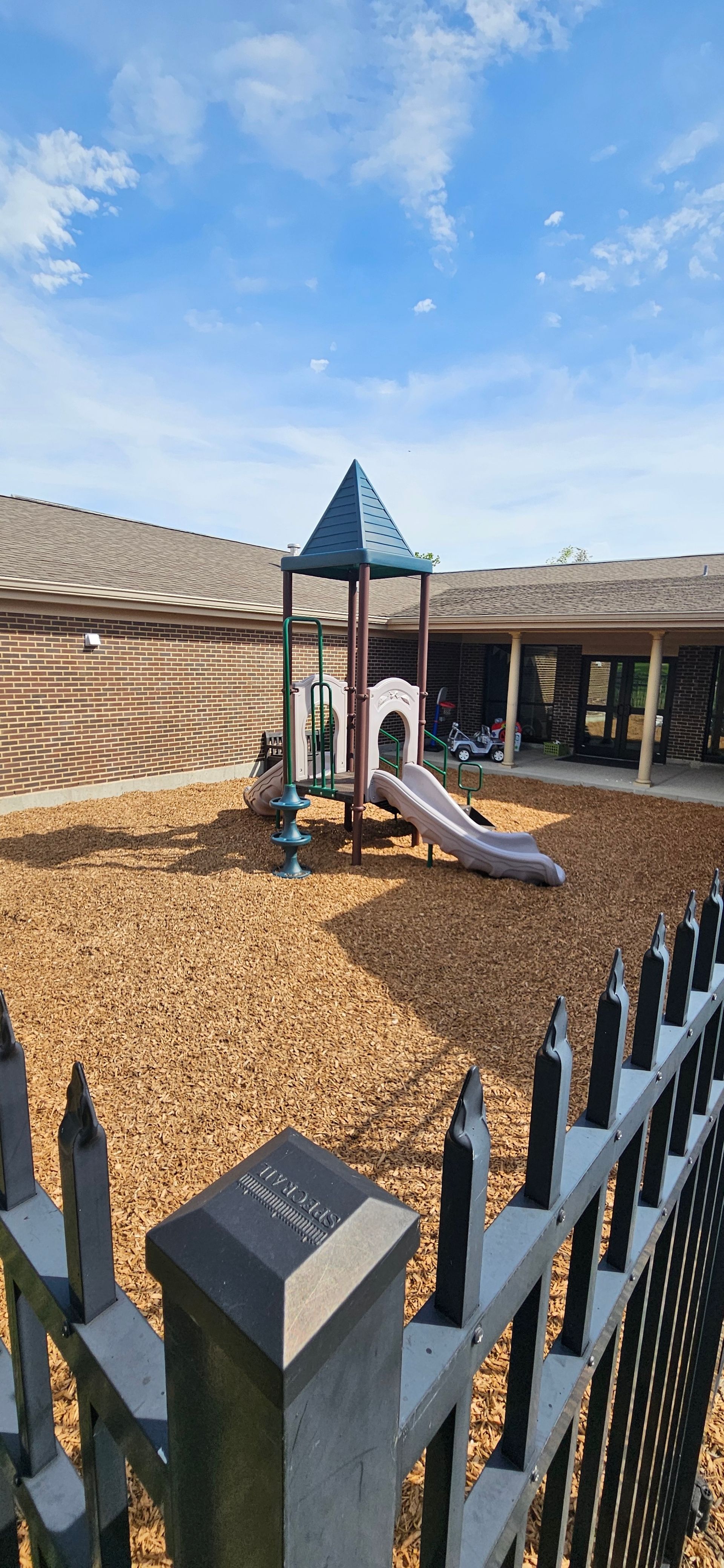 Playground with a slide and wood chips, fenced in. Sunny day, blue sky.