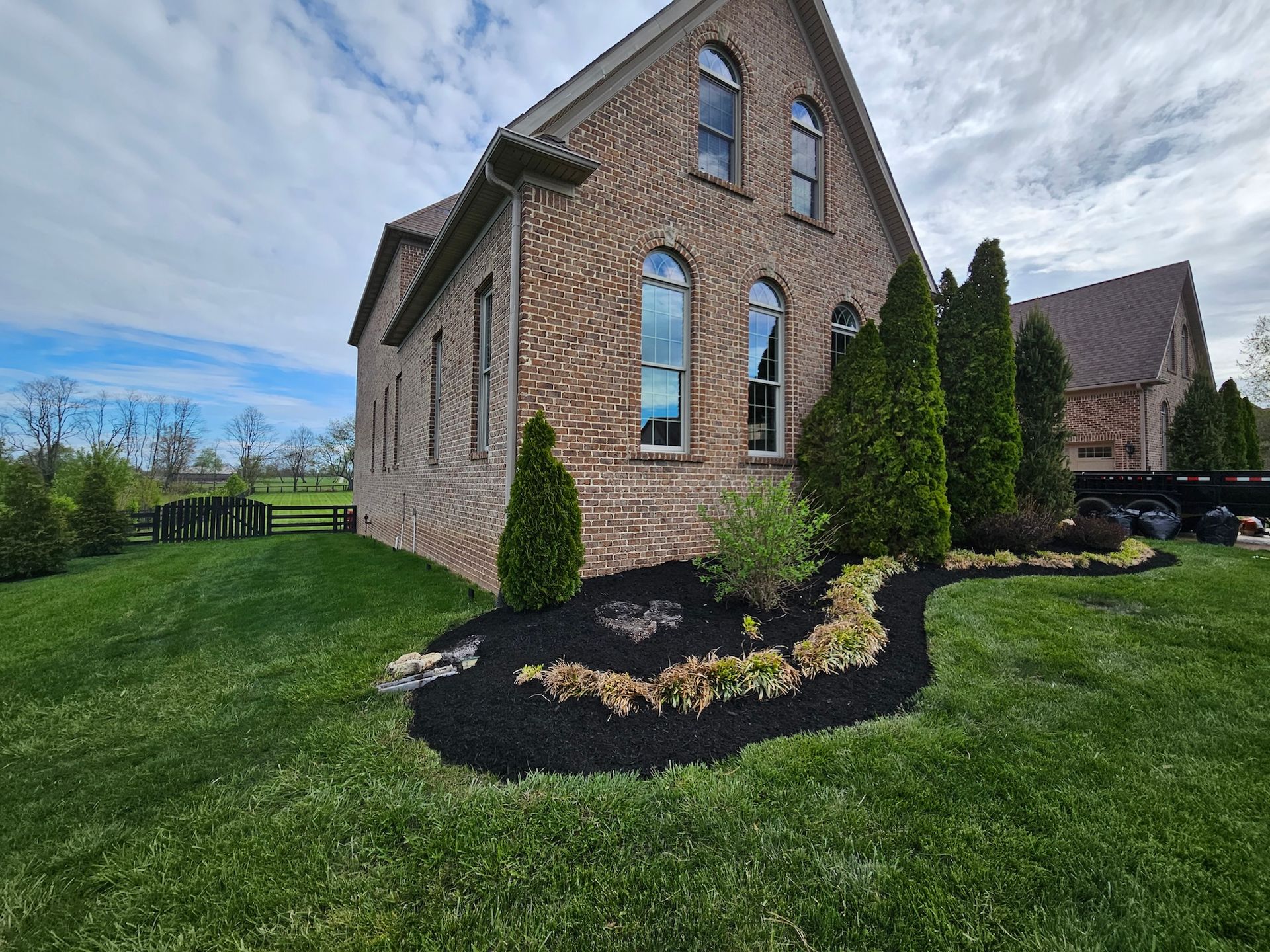Brick house with landscaping, including green lawn, dark mulch, and evergreen trees. Overcast sky.