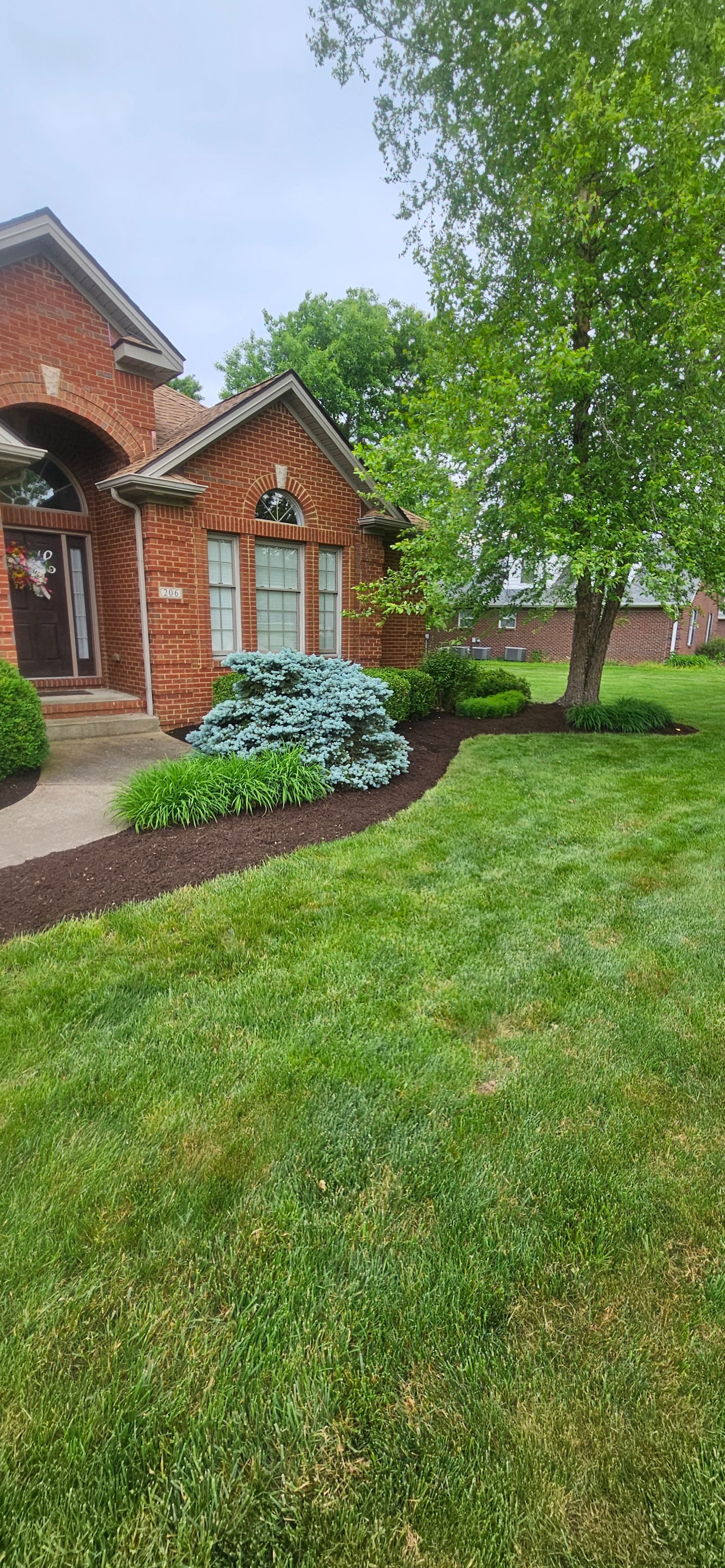 Red brick house with manicured lawn, mulched flower beds, and a large tree.