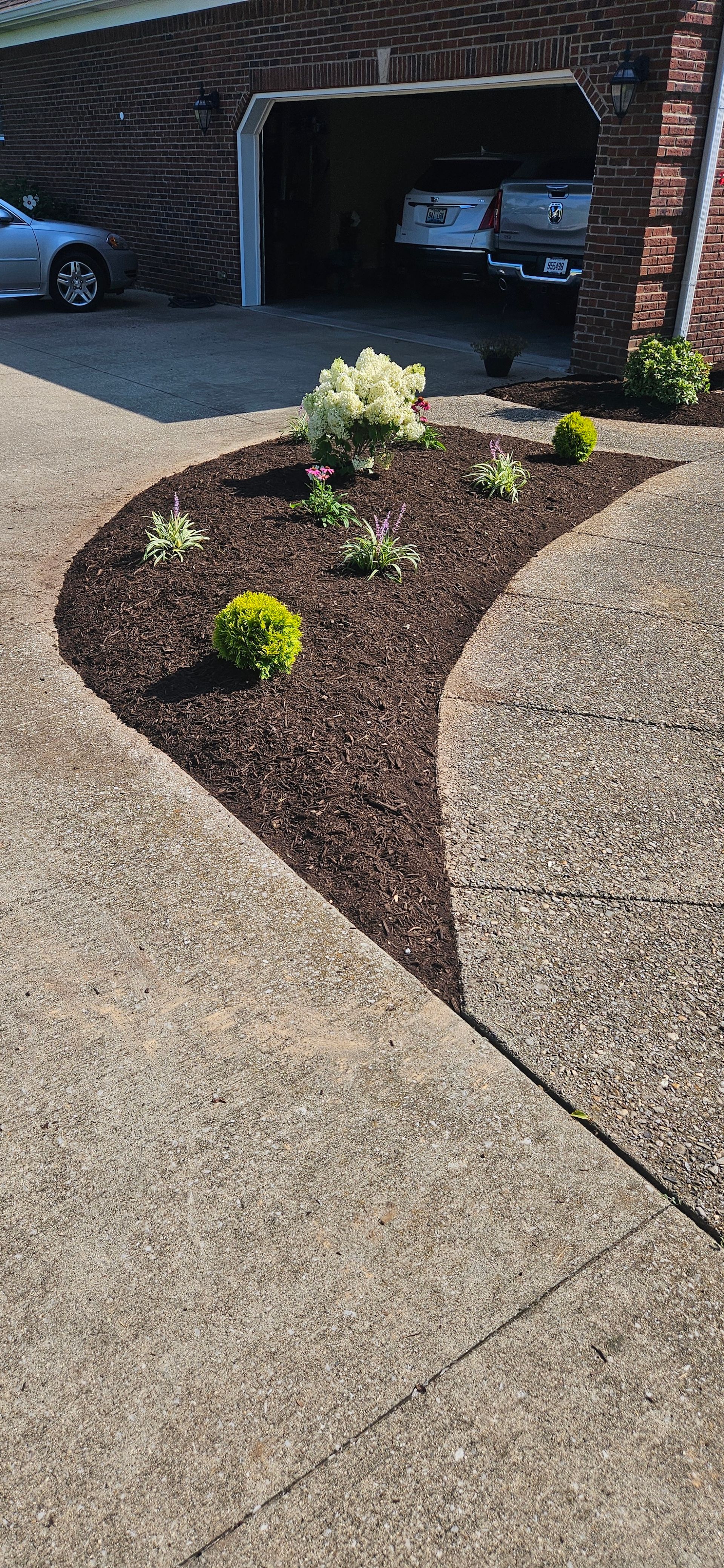 A gravel driveway curves around a garden bed with plants and mulch, in front of a garage.