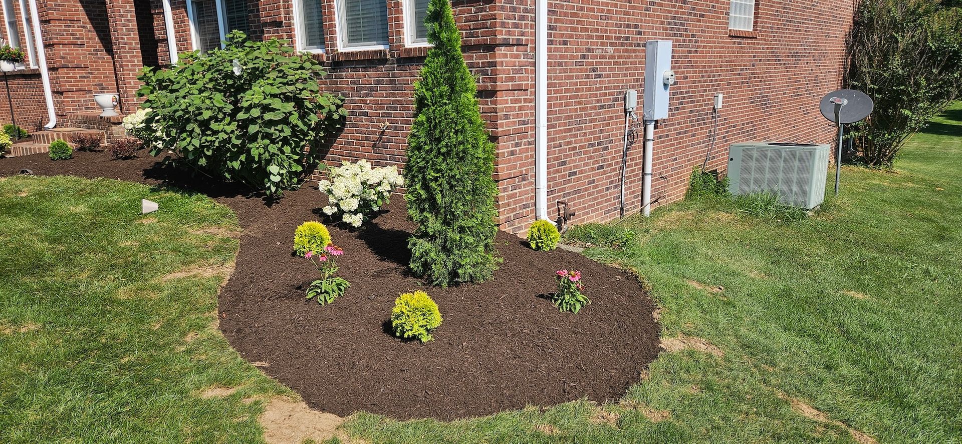 Landscaped flower bed with trees and shrubs in front of a brick building and grass lawn.