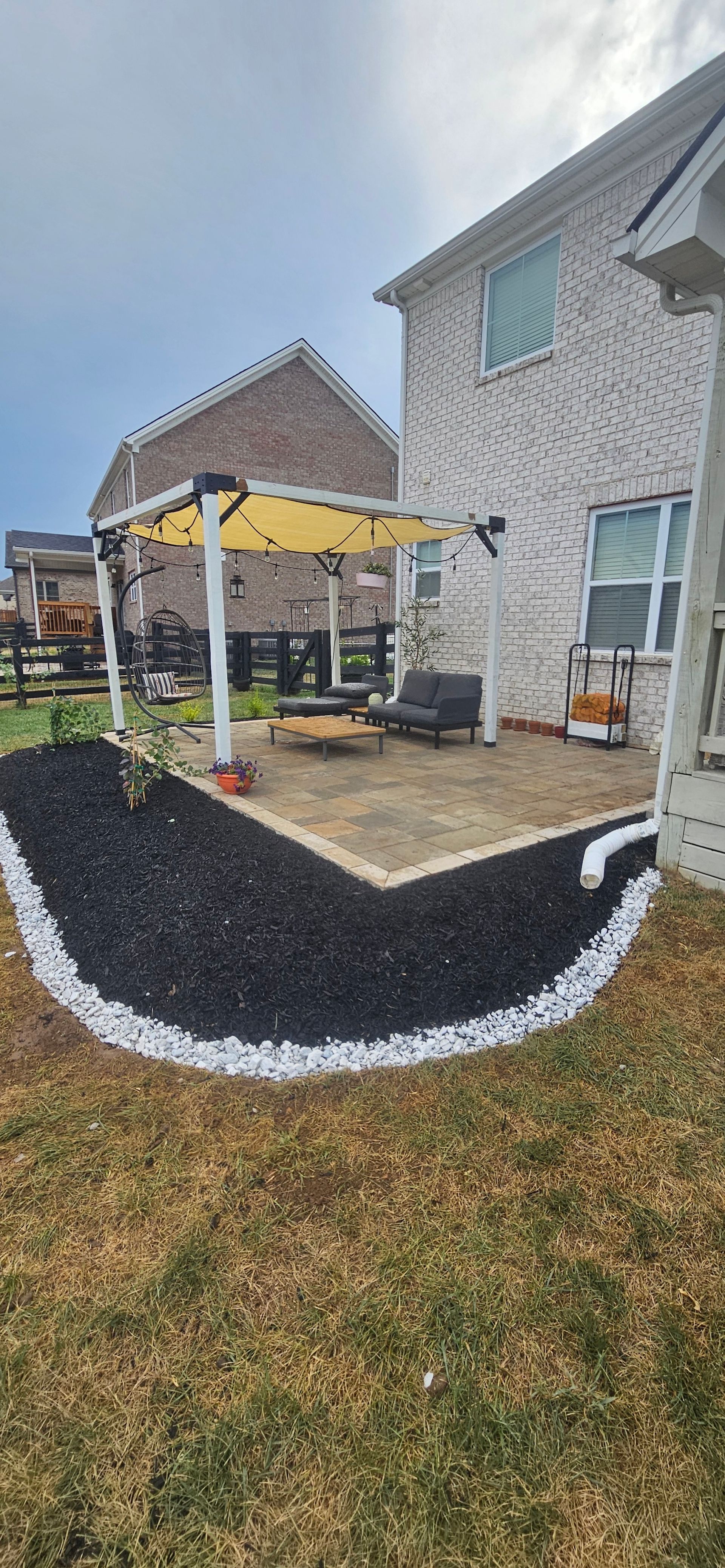 Backyard landscaping with a patio, black mulch, and white border. A pergola with yellow shade, next to a brick house.